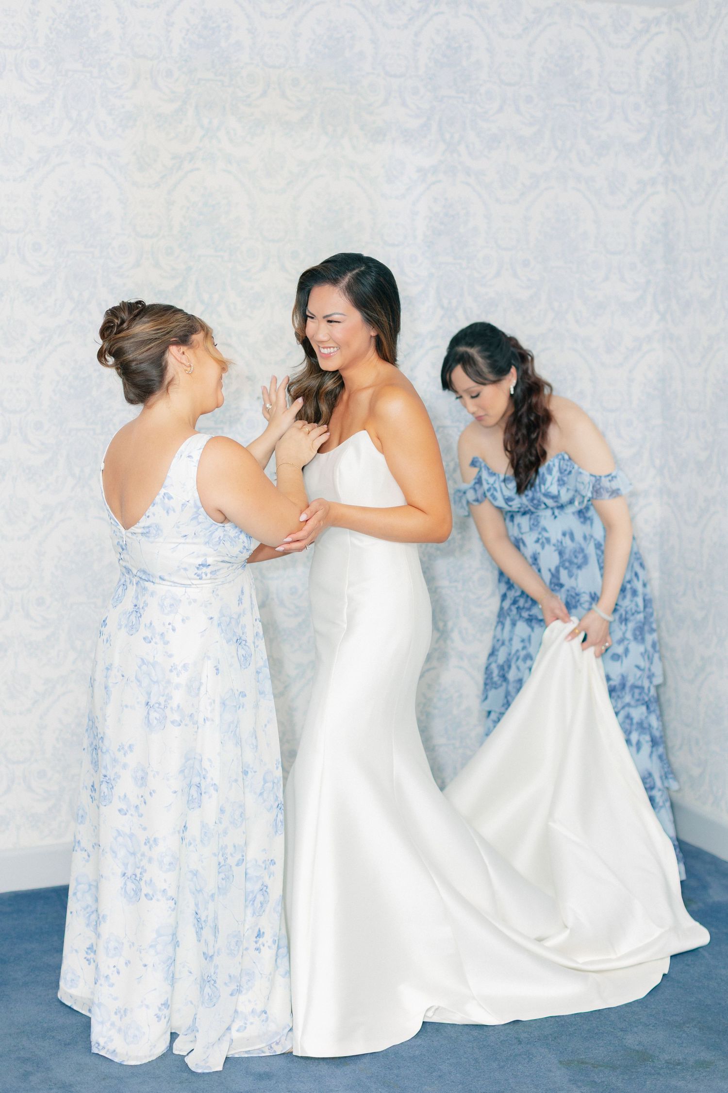 Bride in white gown gets help from two bridesmaids in blue floral dresses while getting ready.