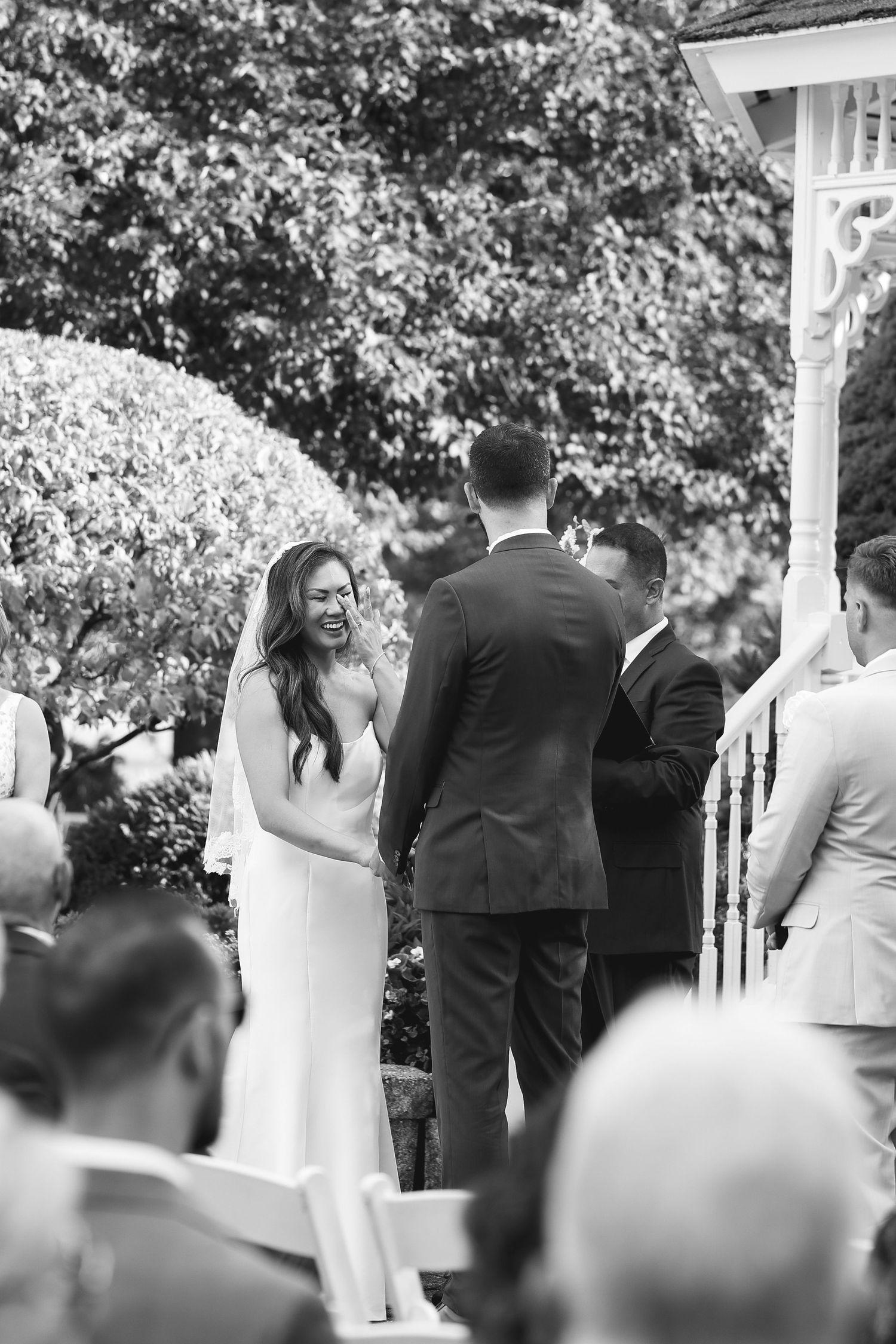Black and white outdoor wedding ceremony with smiling bride in white gown holding hands with groom in suit before guests.