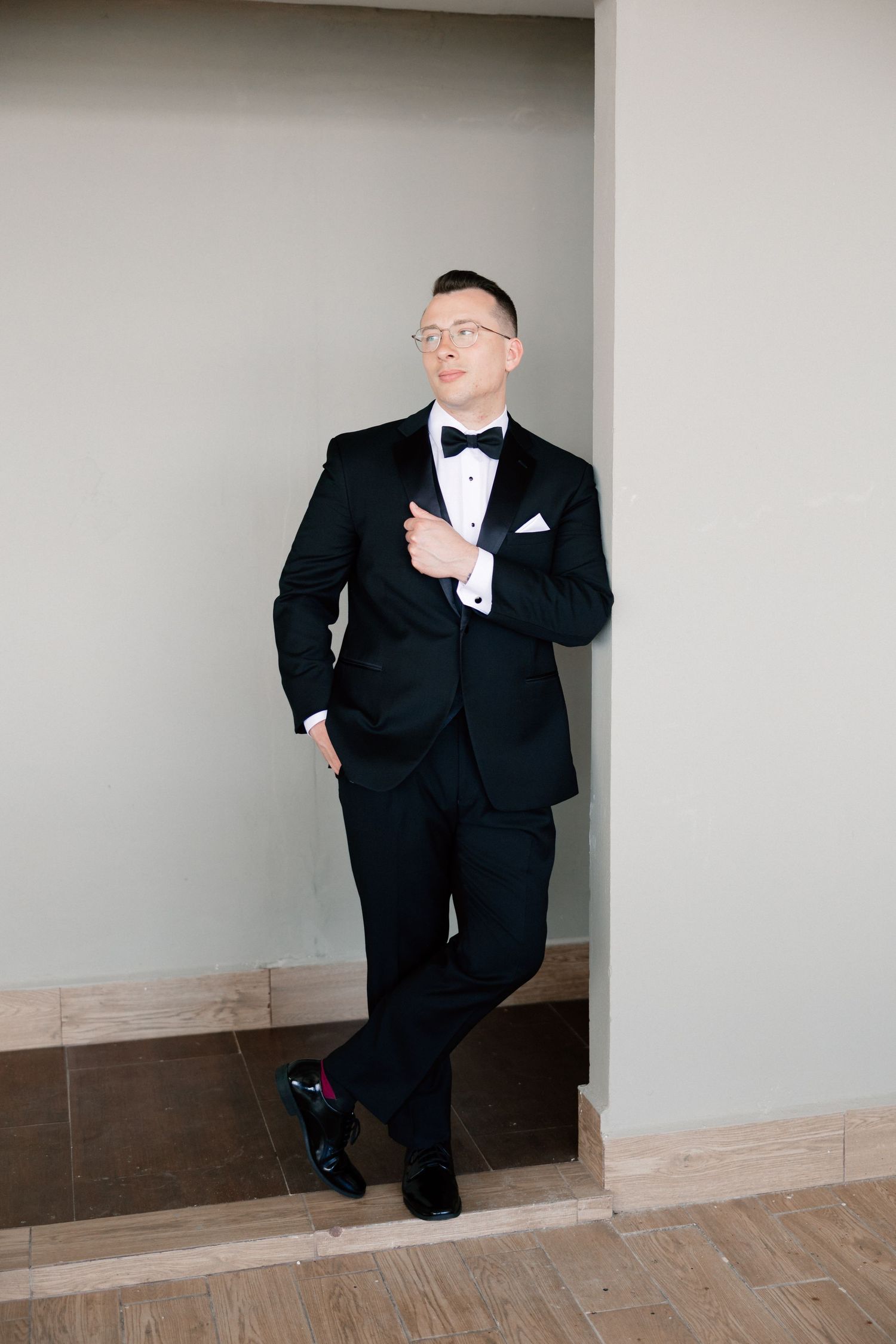 Elegant groom in black tuxedo with bow tie and glasses leaning against wall in formal wedding attire.