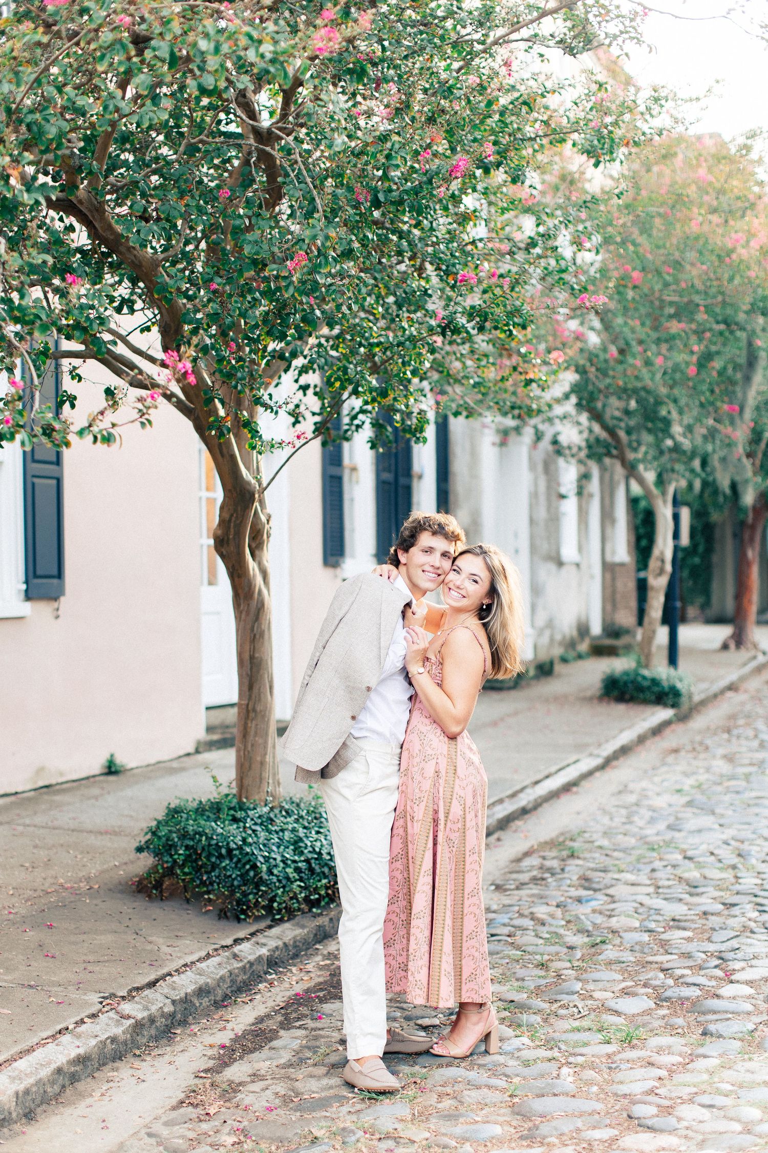 Couple embracing on a charming cobblestone street lined with blooming trees and pastel buildings.