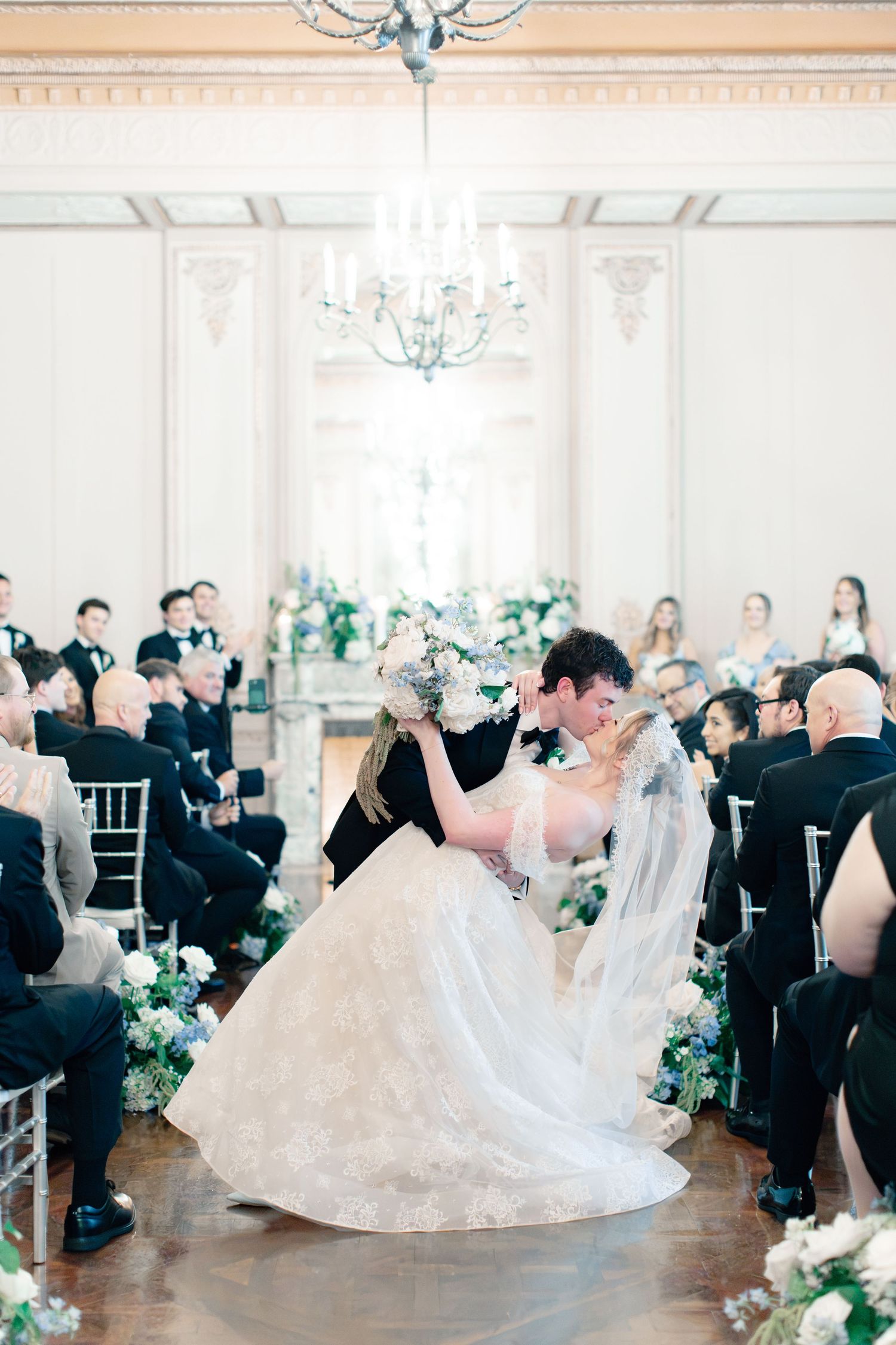Bride and groom share first kiss during elegant ballroom wedding ceremony with chandelier and floral arrangements.