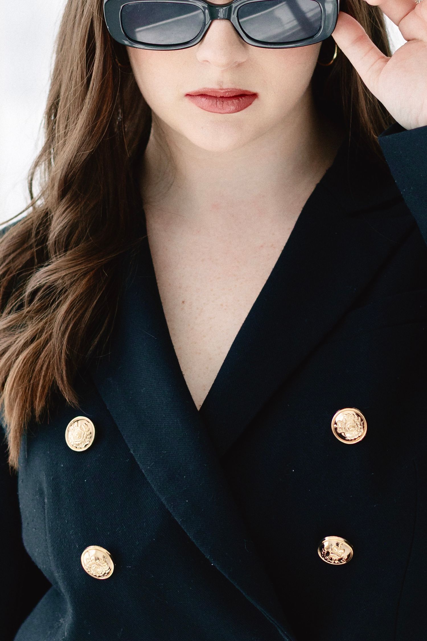 Woman in black double-breasted blazer with gold buttons, holding dark sunglasses, with long brown hair.