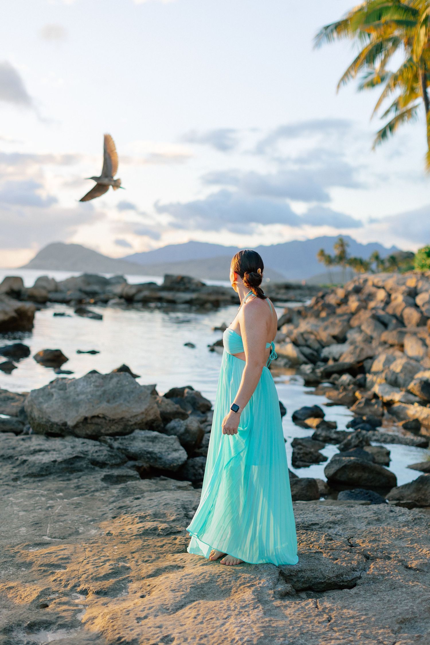 Woman in teal maxi dress standing on rocky Hawaiian beach at sunset with palm tree and bird in sky.