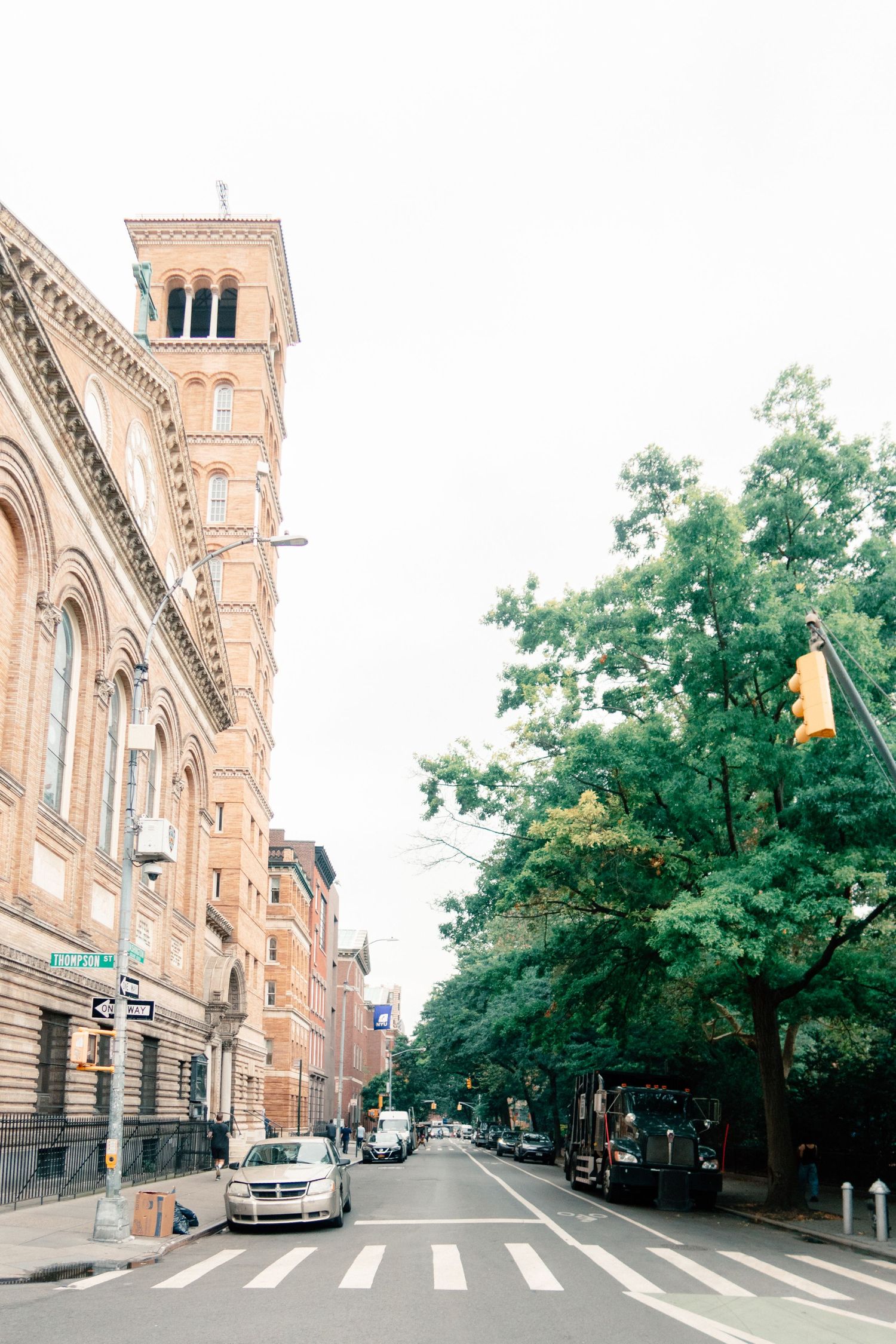 Historic brick church tower on a tree-lined urban street with parked cars and a yellow traffic light.
