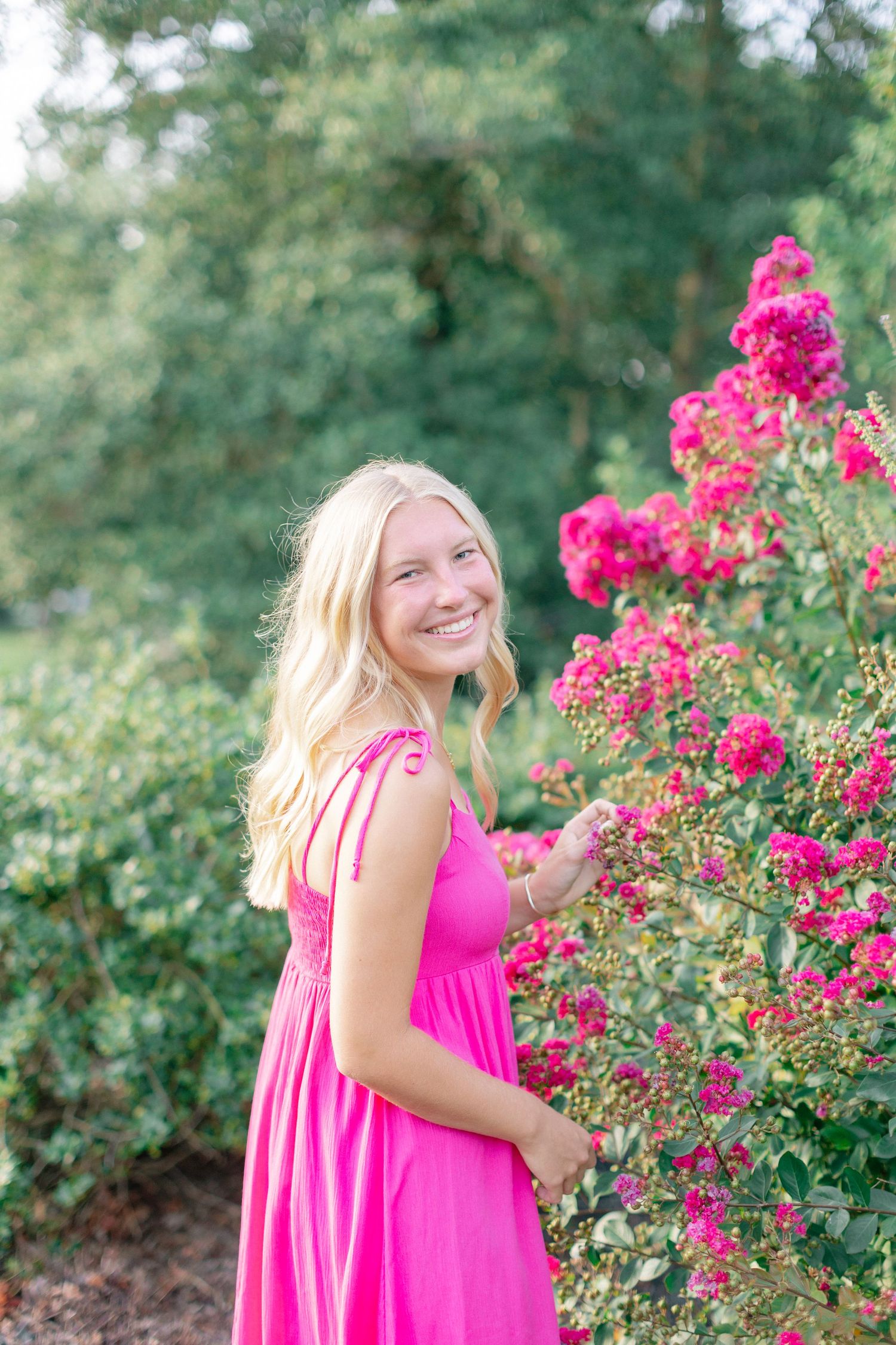 Smiling blonde teen in pink dress touching vibrant pink flowers in lush green garden.