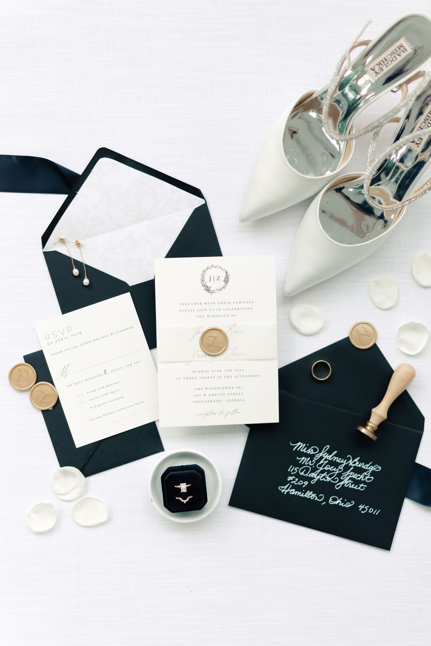 Elegant wedding flat lay with navy envelopes, gold wax seals, white invitations, diamond ring, and bridal heels on white background.