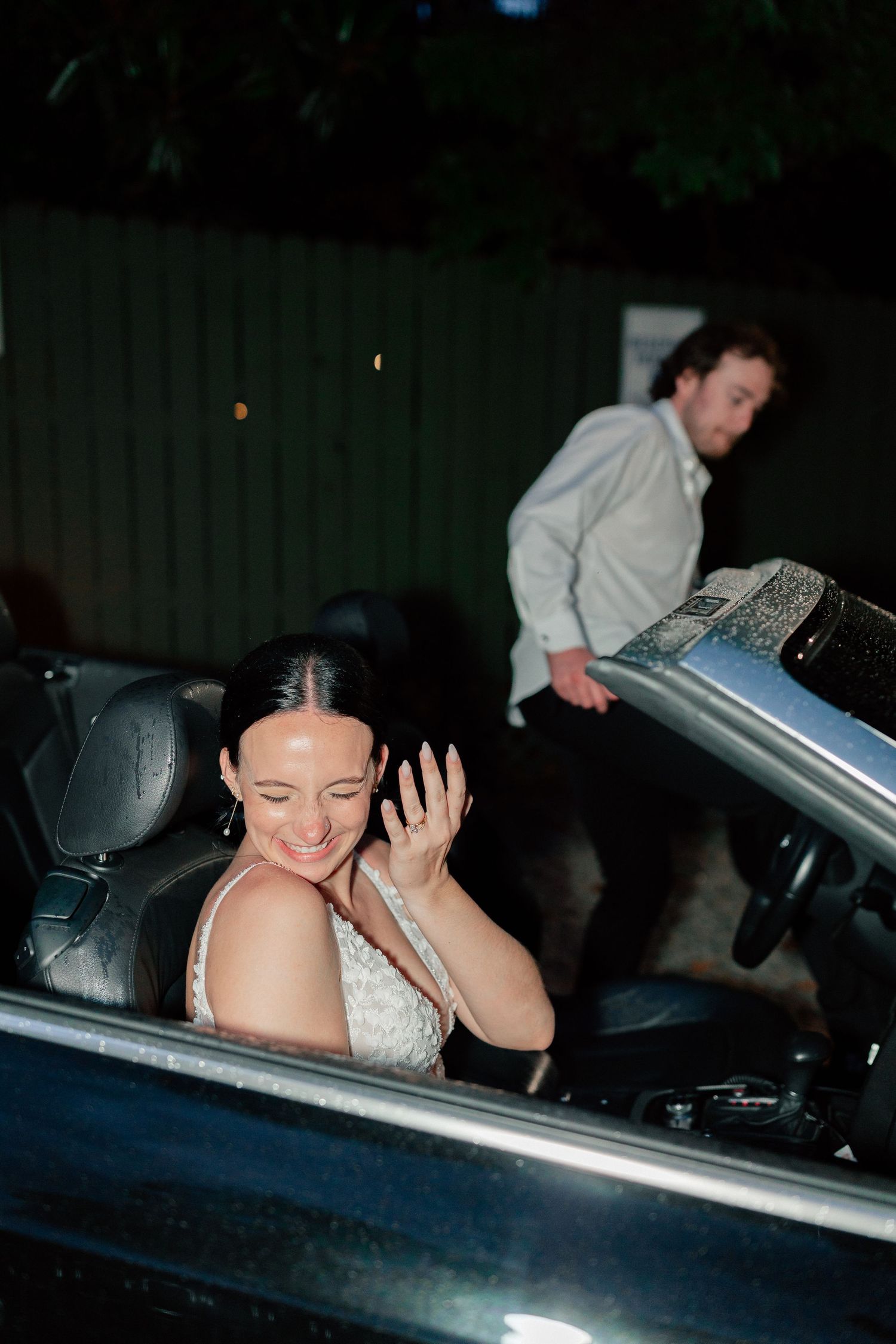 Bride smiling in convertible car at night while groom closes the door behind her at wedding reception.