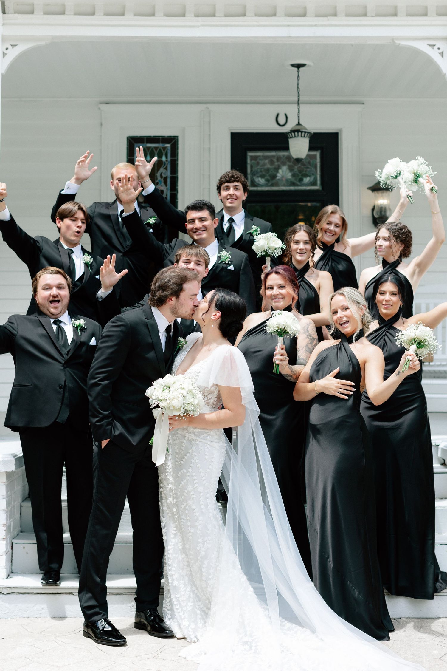 Wedding couple kissing surrounded by bridal party in black formal attire holding white bouquets on white mansion steps.