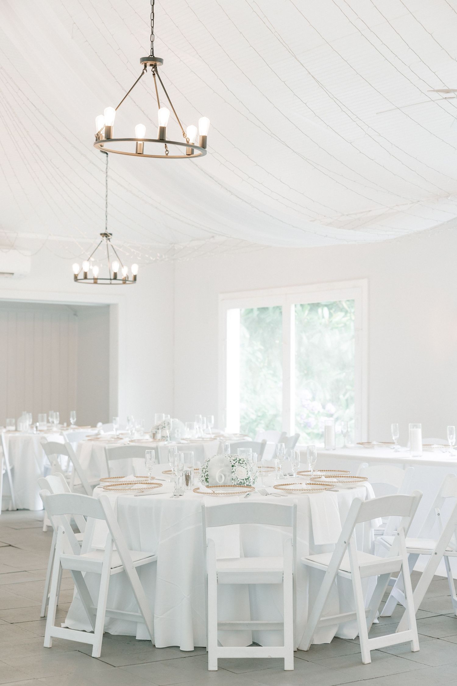 Elegant white wedding reception tent with round tables, white folding chairs, gold chandeliers, and draped ceiling decor.