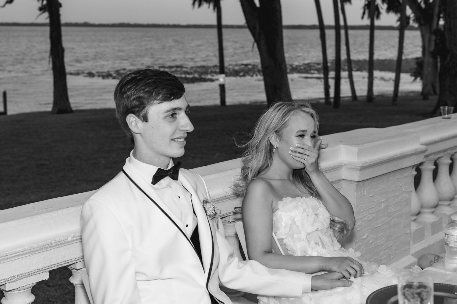 Black and white photo of a young couple in formal wear by waterfront, bride covering mouth in emotional moment, wedding at Bella Cosa