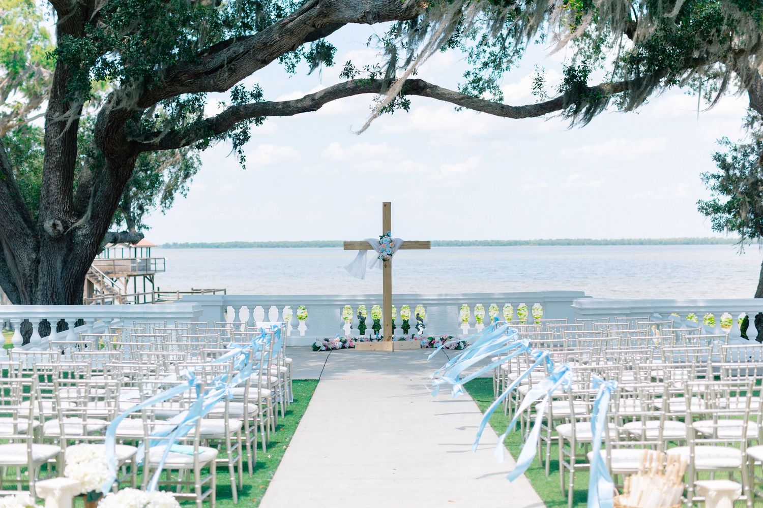 Outdoor waterfront wedding ceremony setup with white chairs, blue ribbons, a wooden cross altar, and a large oak tree by the water, Florida