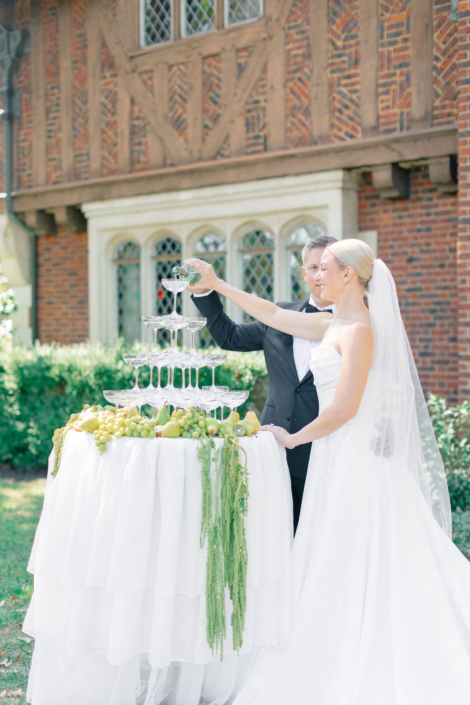 Bride and groom pouring champagne tower surrounded by green grapes and hanging amaranthus at outdoor reception.