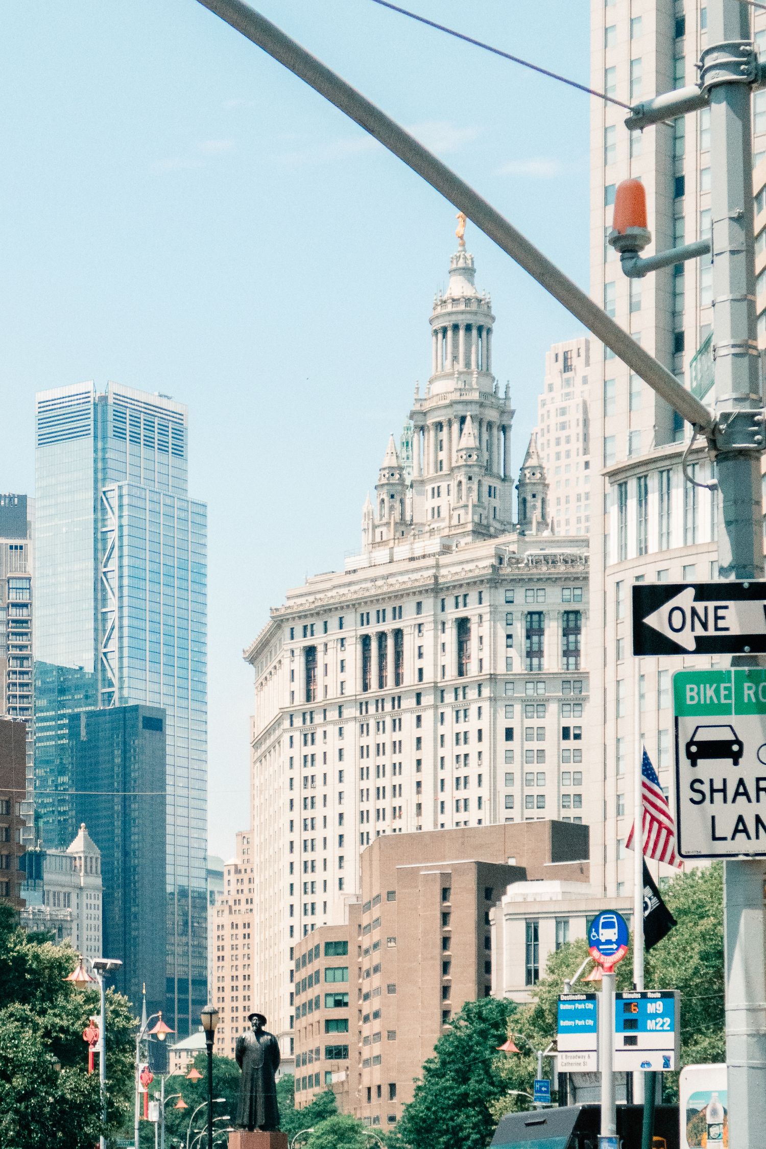 Manhattan Municipal Building rises above NYC street with signs, skyscrapers, and a statue under a clear blue sky.