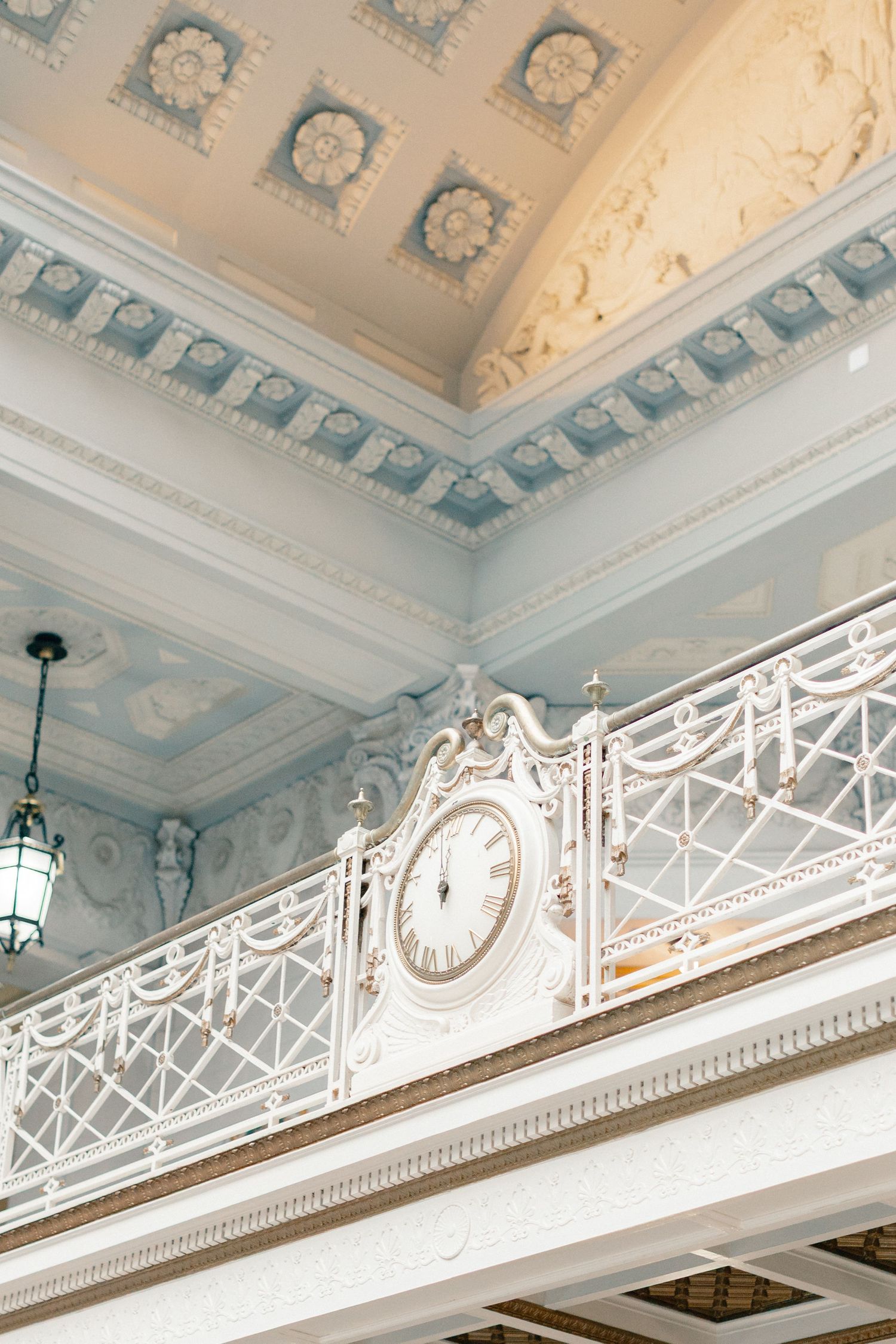 Ornate white balcony with decorative clock, intricate railings, and elegant blue coffered ceiling in a classical interior.