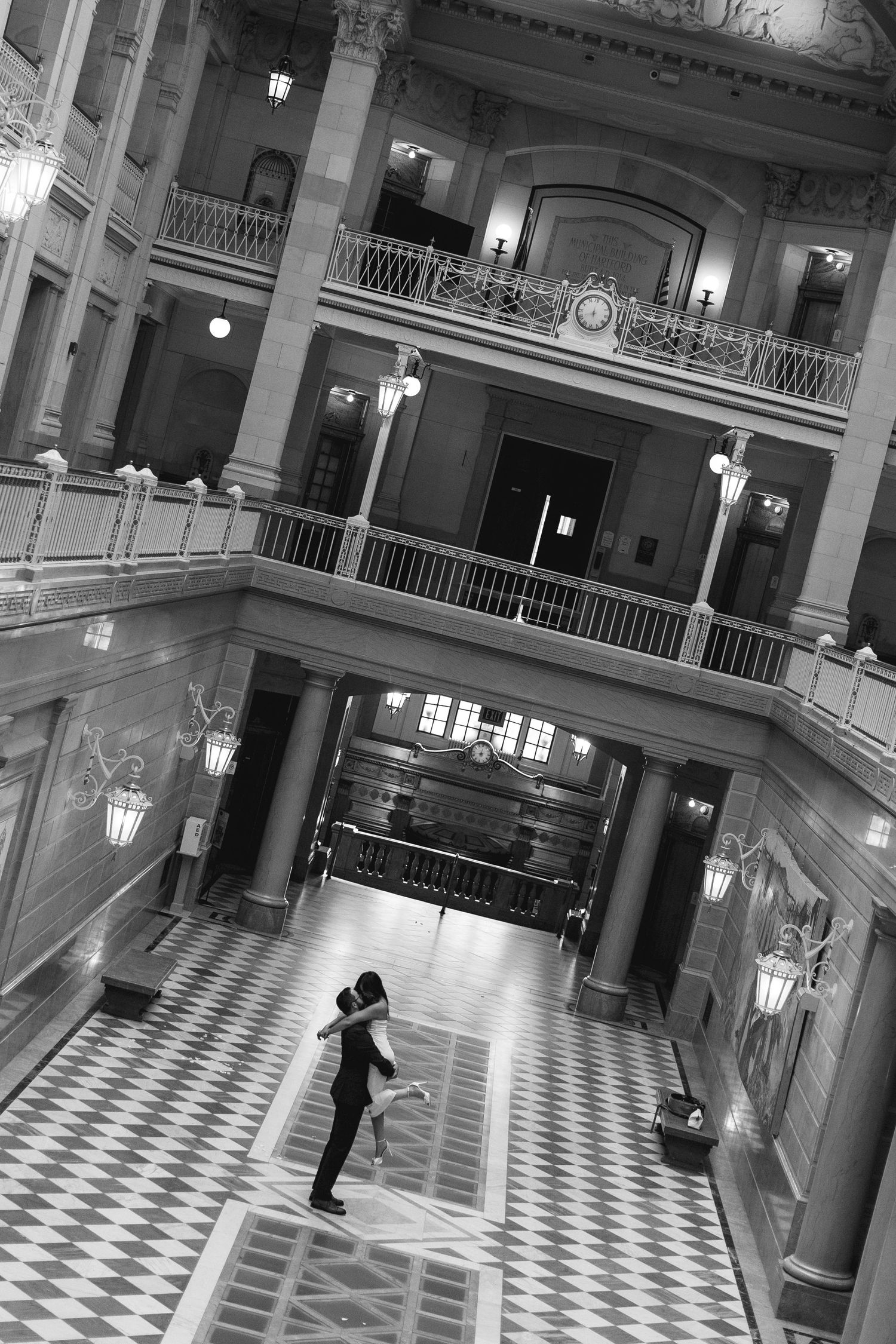 Couple dancing in grand historic building atrium with ornate balconies, columns, and checkered floors in black and white.