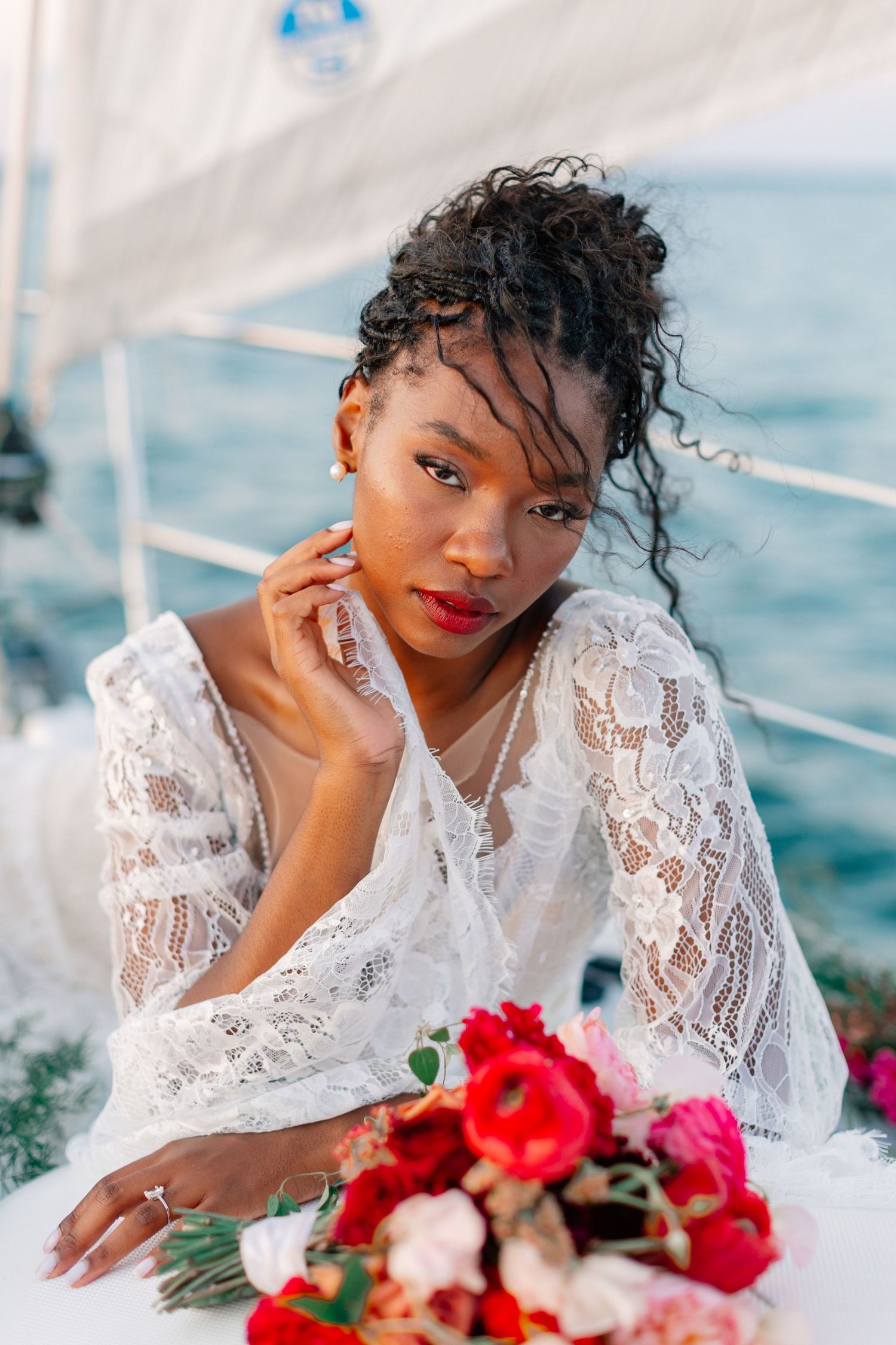 Black bride in white lace dress poses with red bouquet on sailboat deck over blue ocean waters.