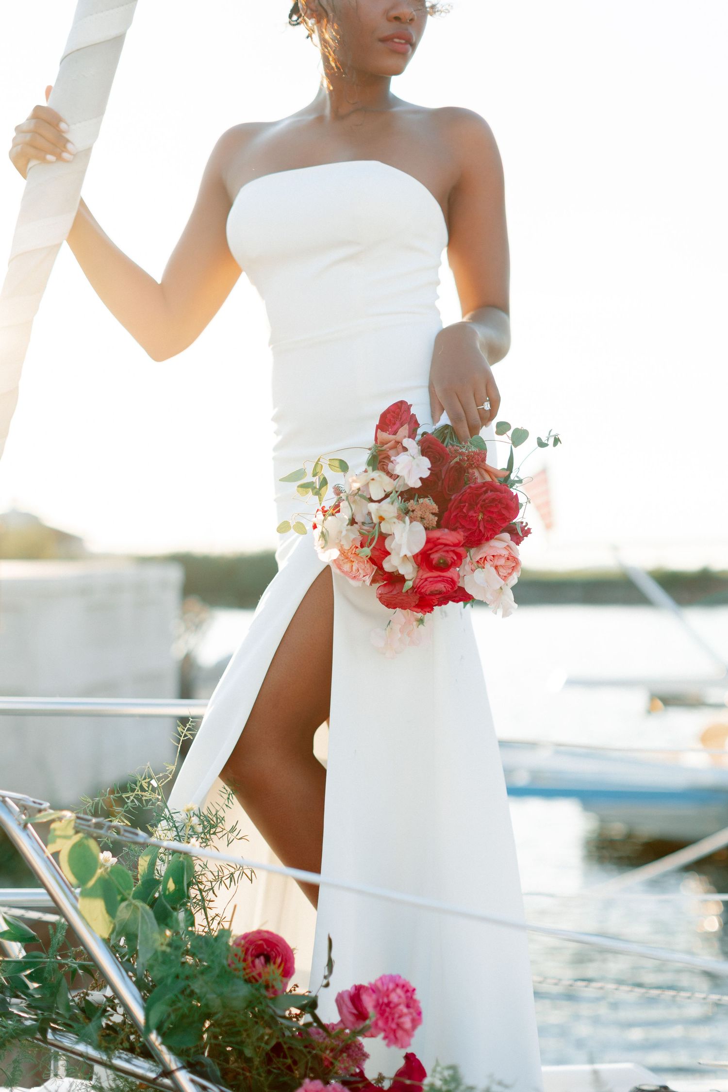 Bride in strapless white gown holding red and pink bouquet on a yacht at golden hour.