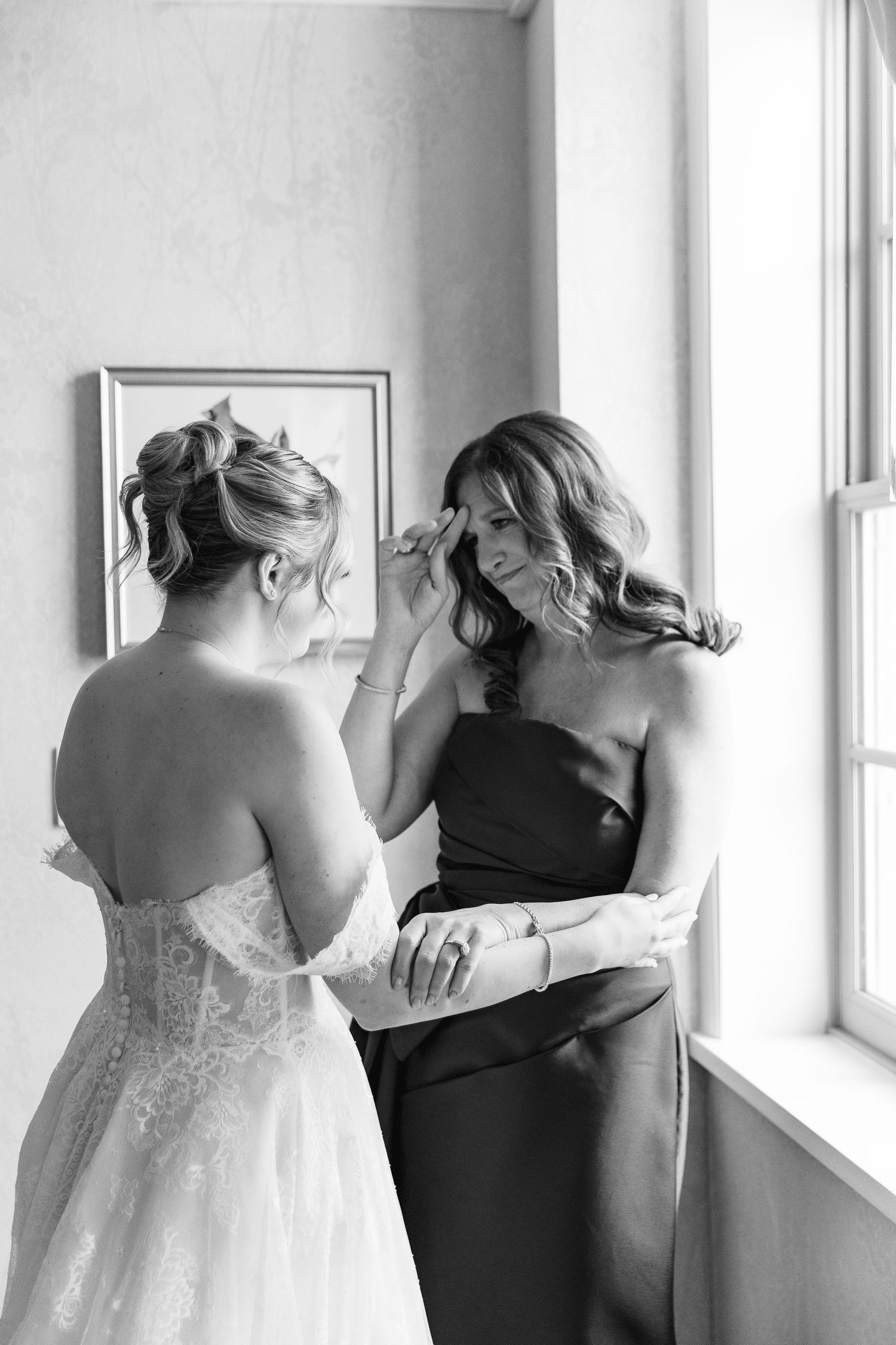 Bride in lace gown gently wipes tear from bridesmaid in strapless dress near window in black and white photo.