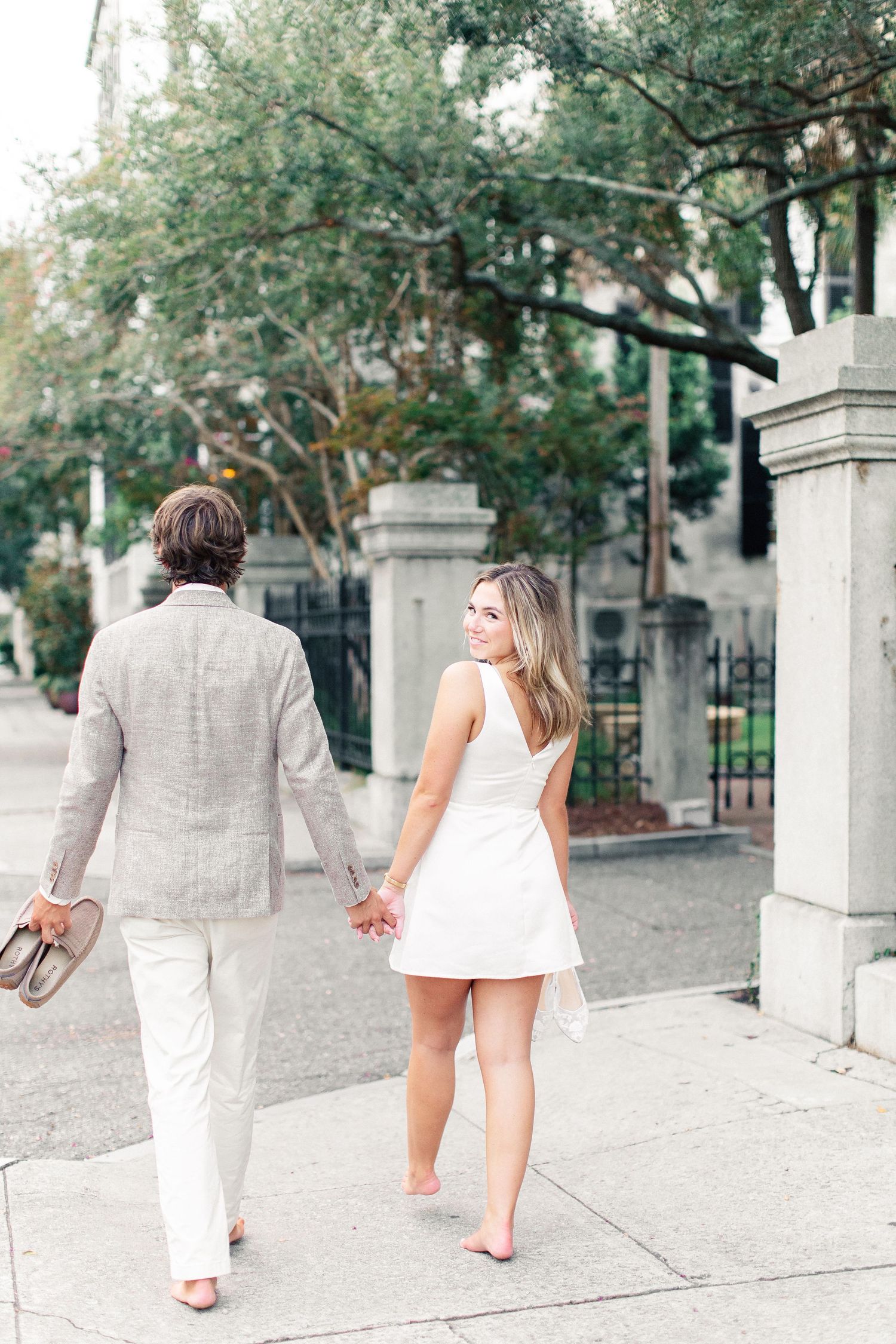 Couple walking hand-in-hand on sidewalk, woman in white mini dress barefoot, man in linen suit carrying shoes.