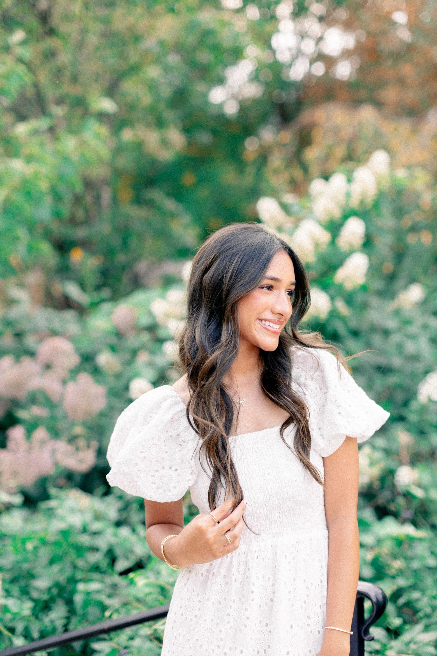 Smiling woman in white eyelet puff-sleeve dress poses outdoors among lush green and white flowering shrubs.