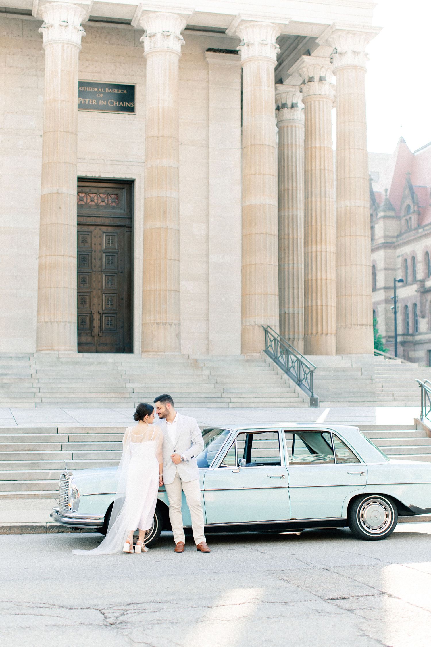 Bride and groom pose beside a vintage mint Mercedes outside a grand columned building on their wedding day.