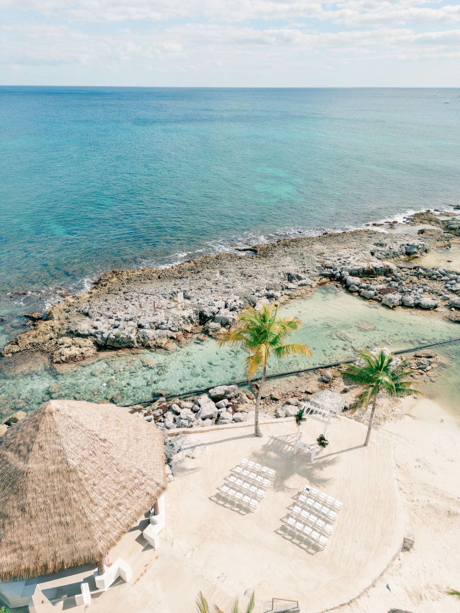 Aerial view of a tropical resort beach with palm trees, lounge chairs, thatched hut, and crystal-clear turquoise ocean waters.