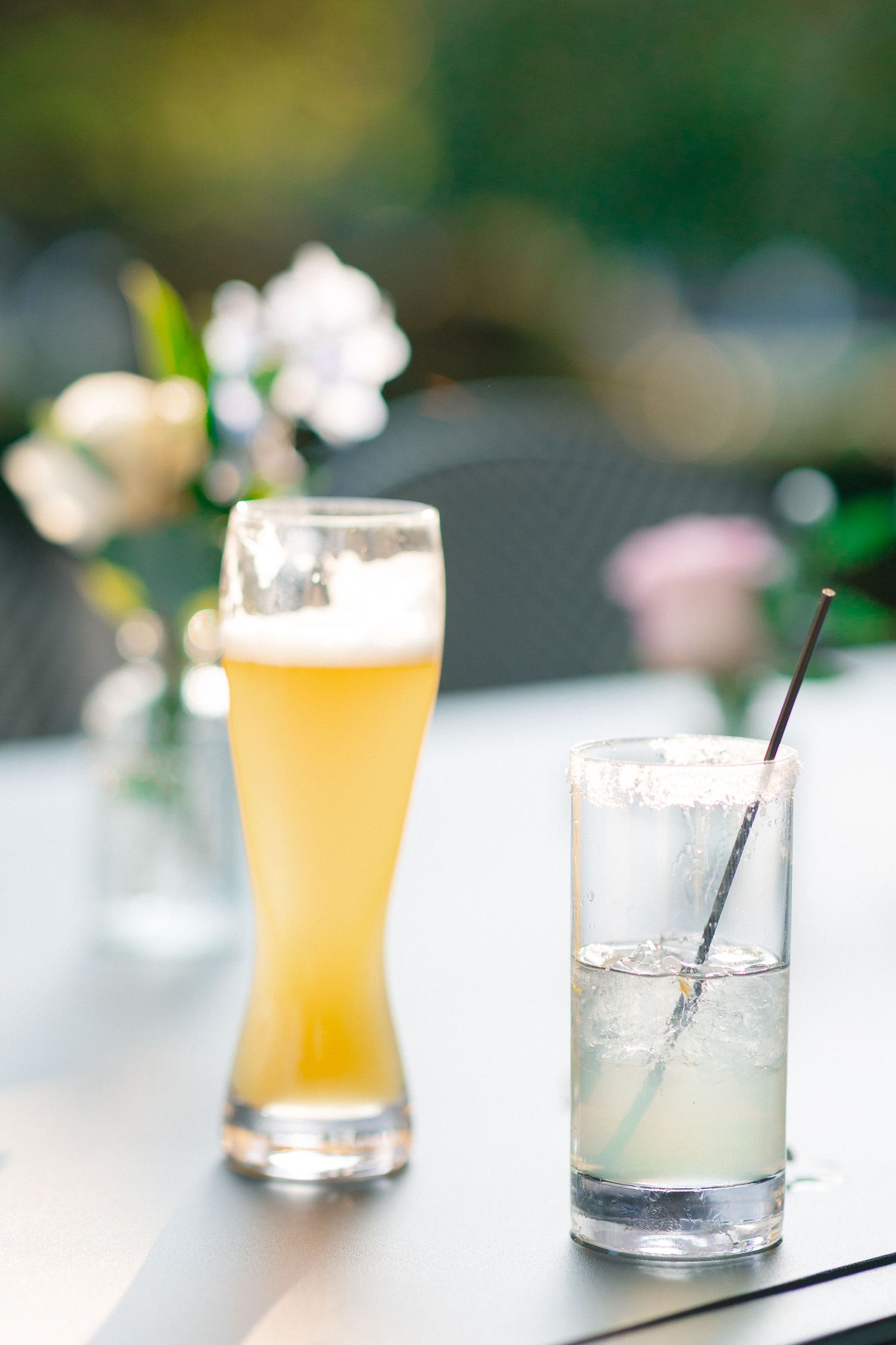 Two refreshing drinks on outdoor table, a wheat beer and iced lemonade with straw, with flowers in background.