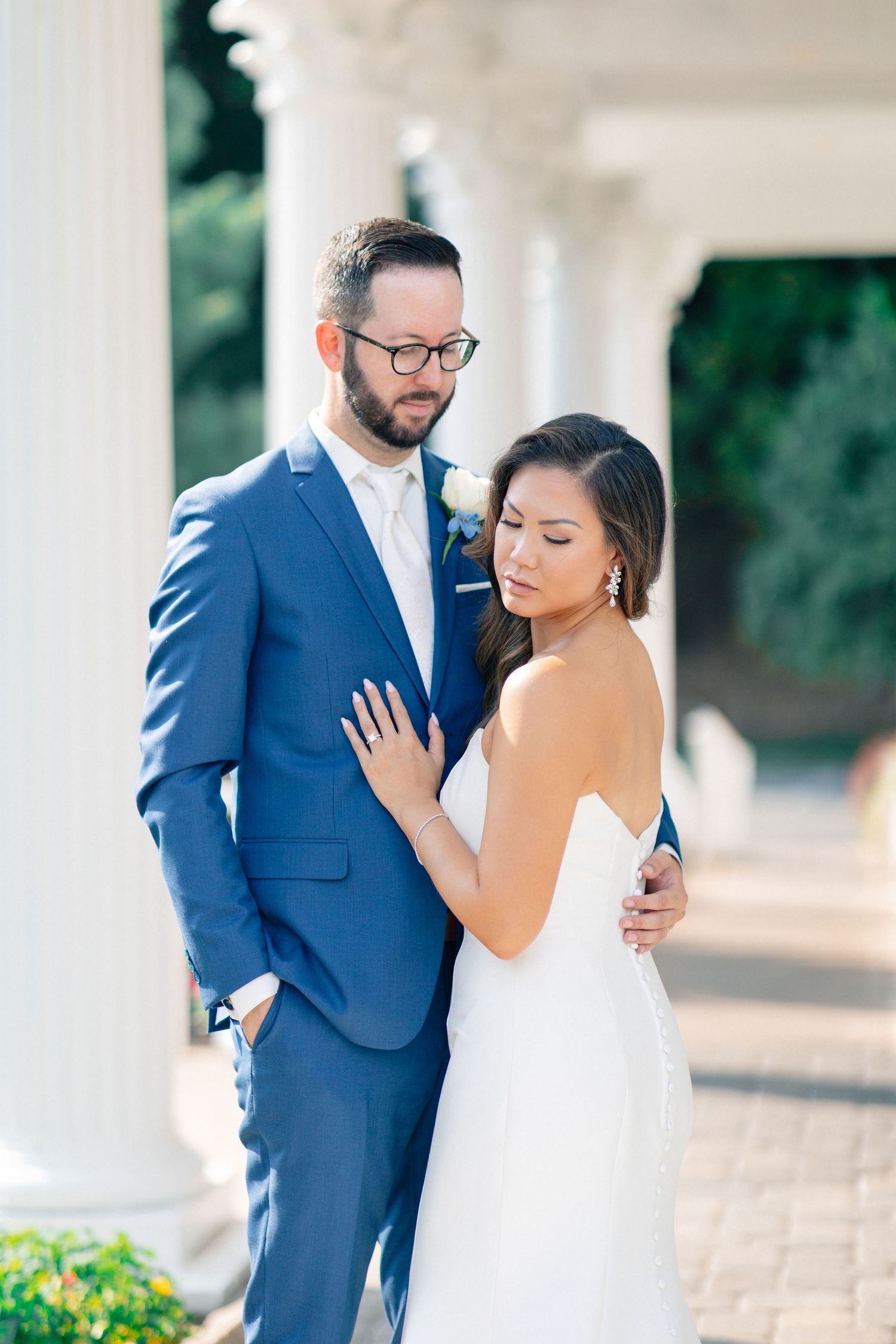 Bride and groom pose elegantly by white columns, groom in navy suit, bride in strapless white gown.