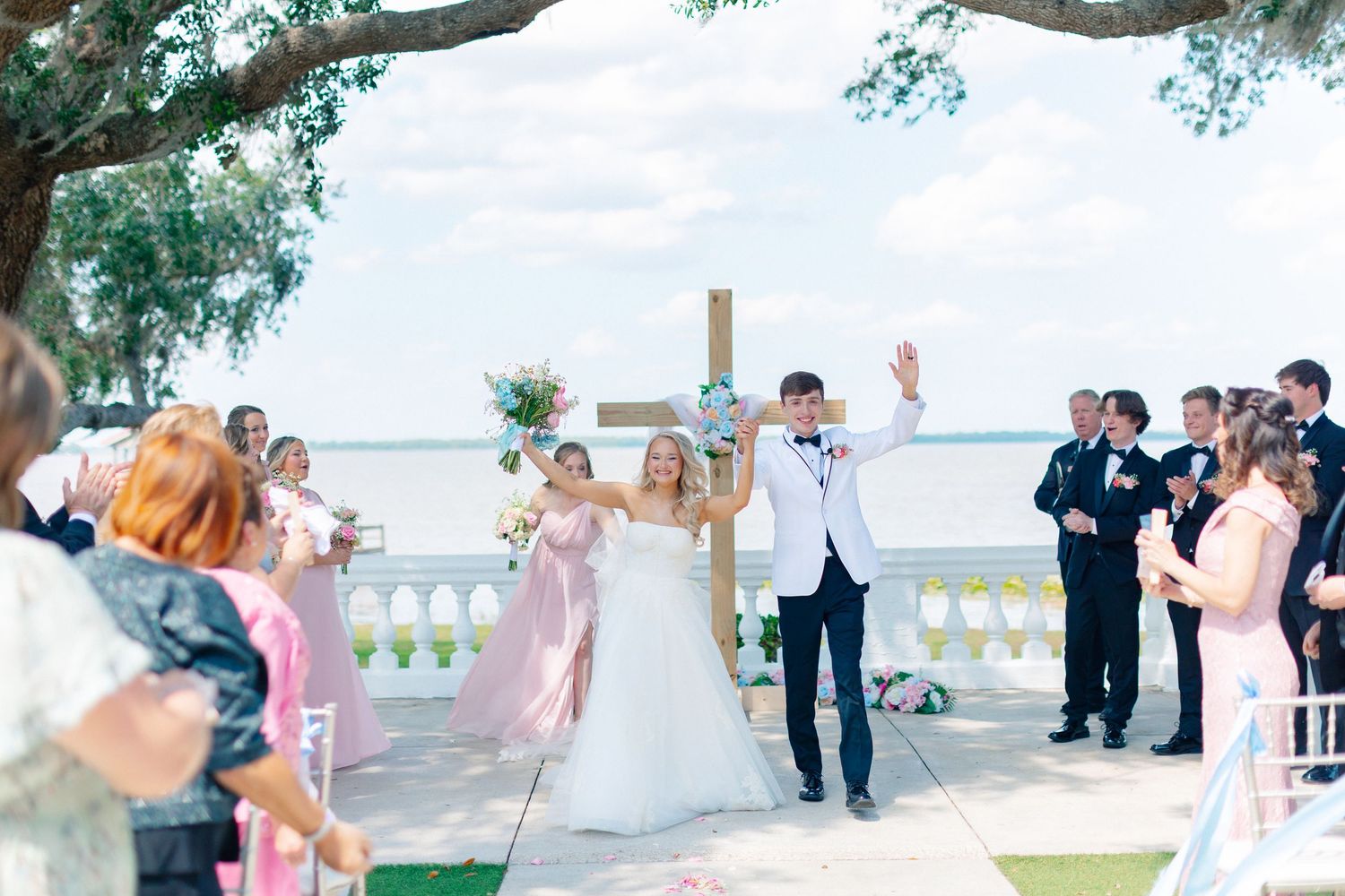 Joyful bride and groom celebrate after outdoor waterfront wedding ceremony with cross backdrop and guests cheering, wedding at Bella Cosa