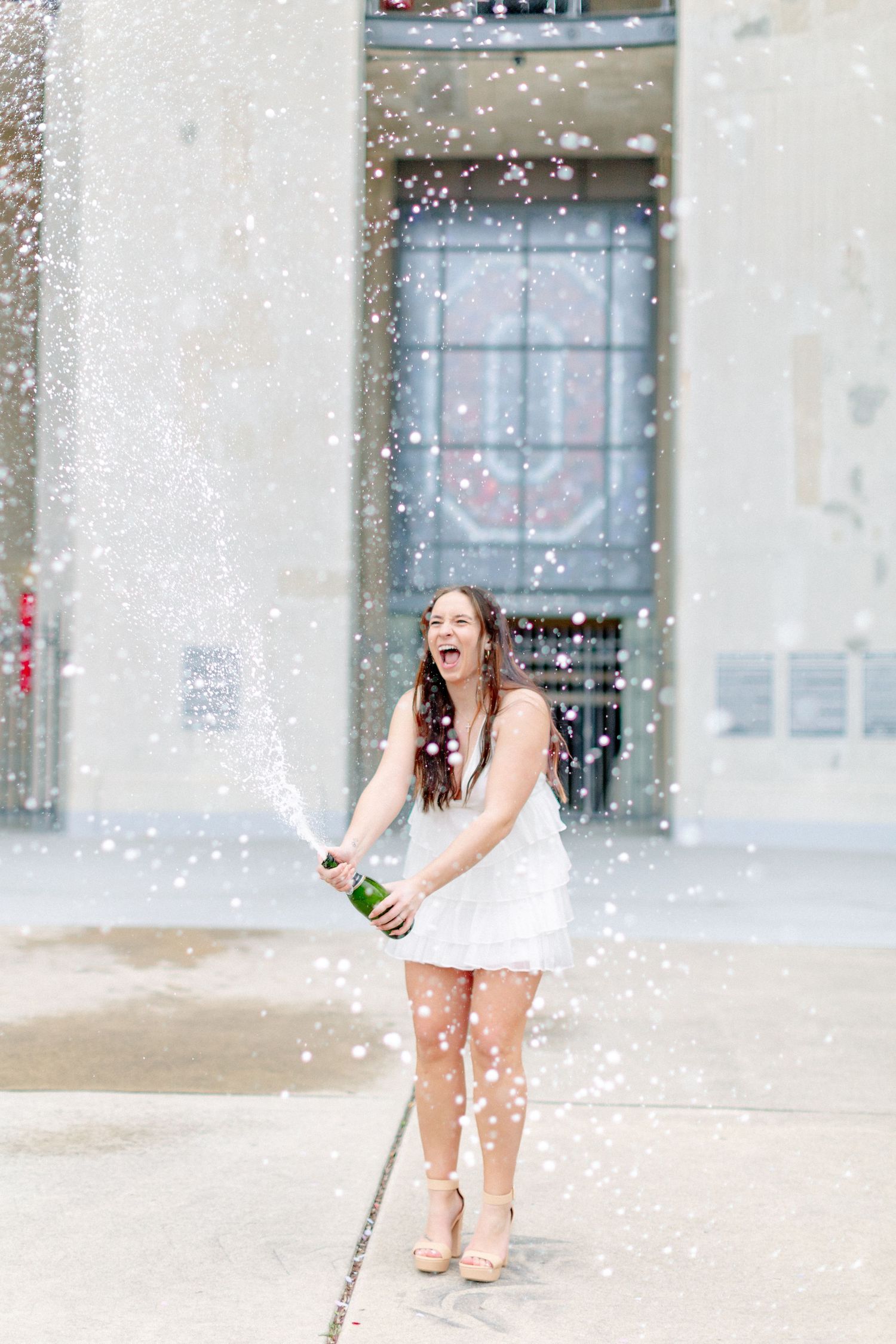 A laughing young woman in a white dress pops a champagne bottle outdoors, sending a festive spray into the air at Ohio State