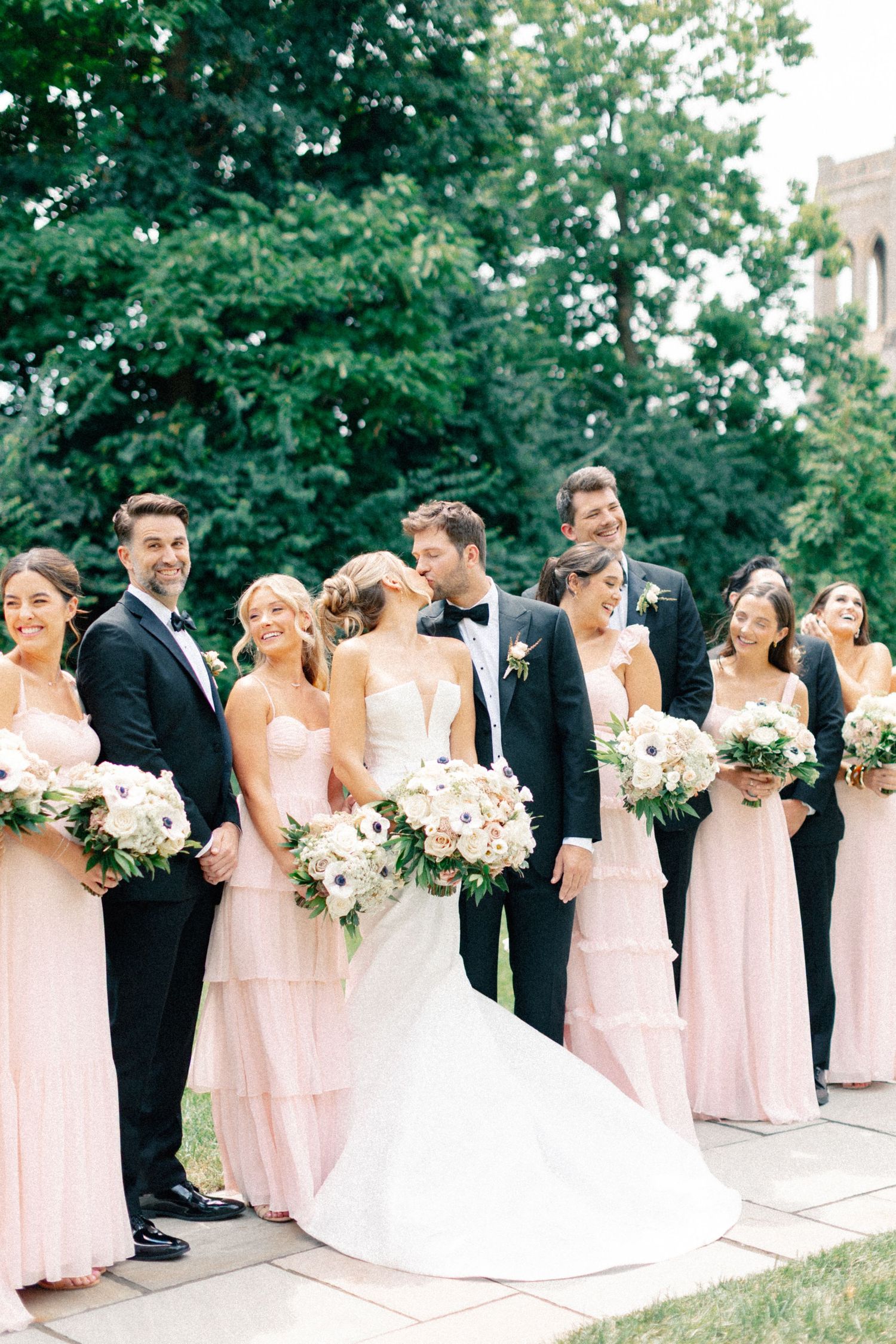 Wedding party poses outdoors, bridesmaids in blush pink gowns holding white bouquets, groomsmen in black tuxedos.
