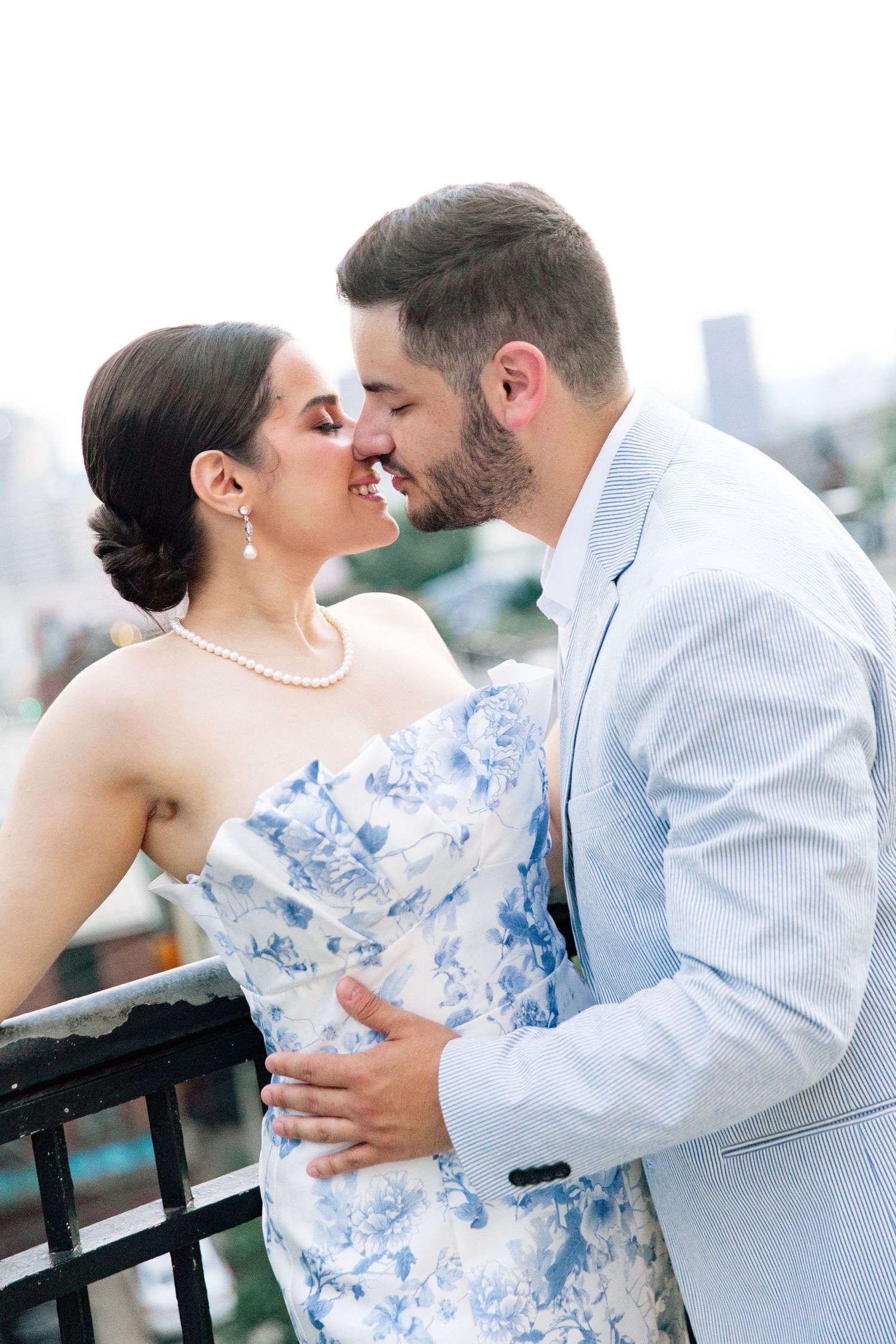 Couple sharing romantic kiss on rooftop, bride in blue floral dress with pearl necklace and groom in light suit.
