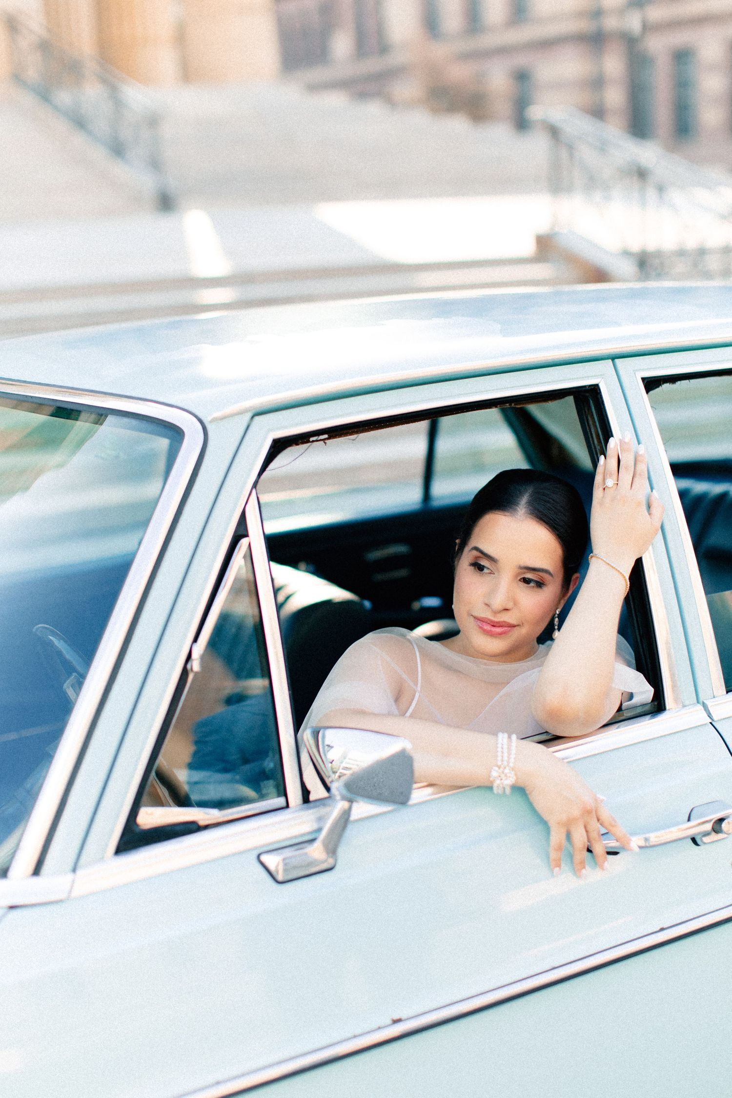 Elegant woman in sheer top leaning out window of vintage light blue classic car on city street.