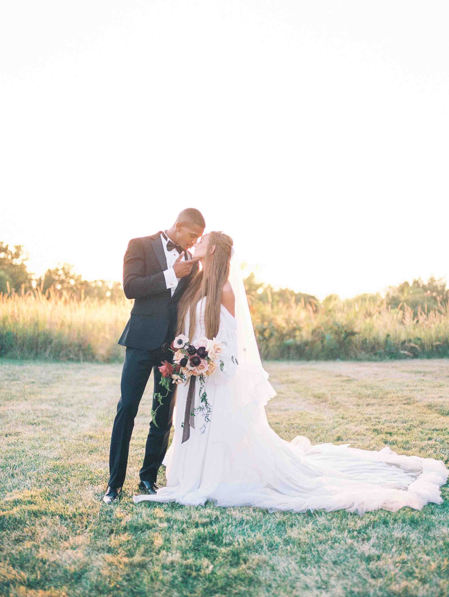 Bride and groom share a romantic kiss at golden hour in a field, with flowing white wedding gown and bouquet.