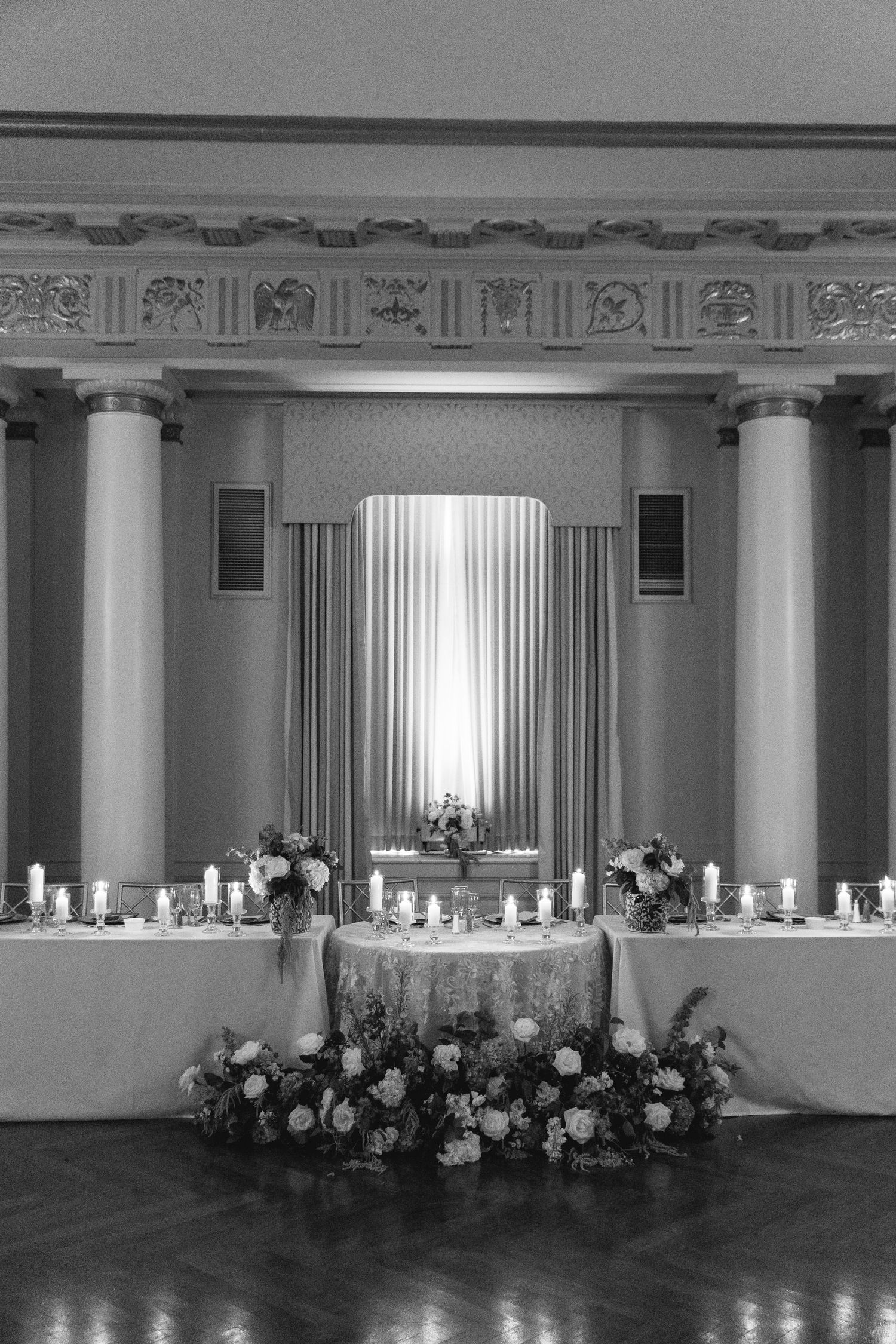 Black and white photo of an elegant wedding reception head table adorned with candles and floral arrangements in a grand ballroom.