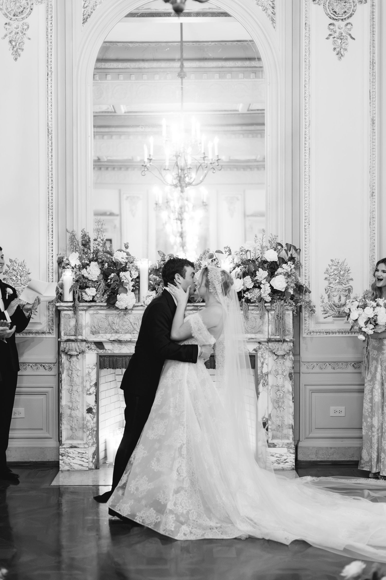 Black and white photo of bride and groom sharing first kiss at elegant indoor wedding ceremony with chandelier.