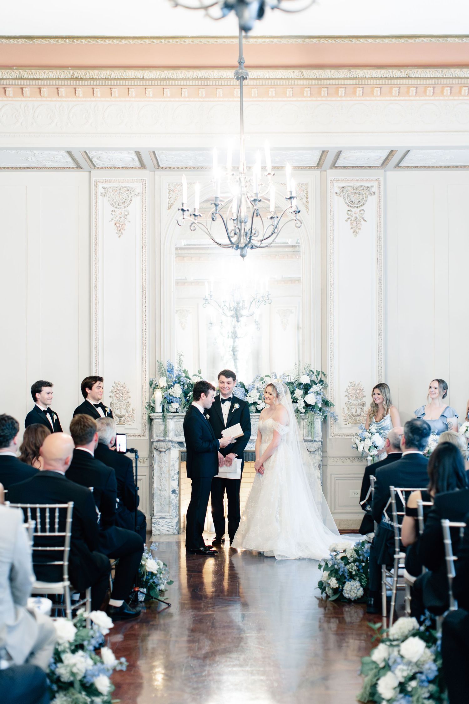 Elegant indoor wedding ceremony in a grand ballroom with chandelier, floral arrangements, and couple exchanging vows at the altar.