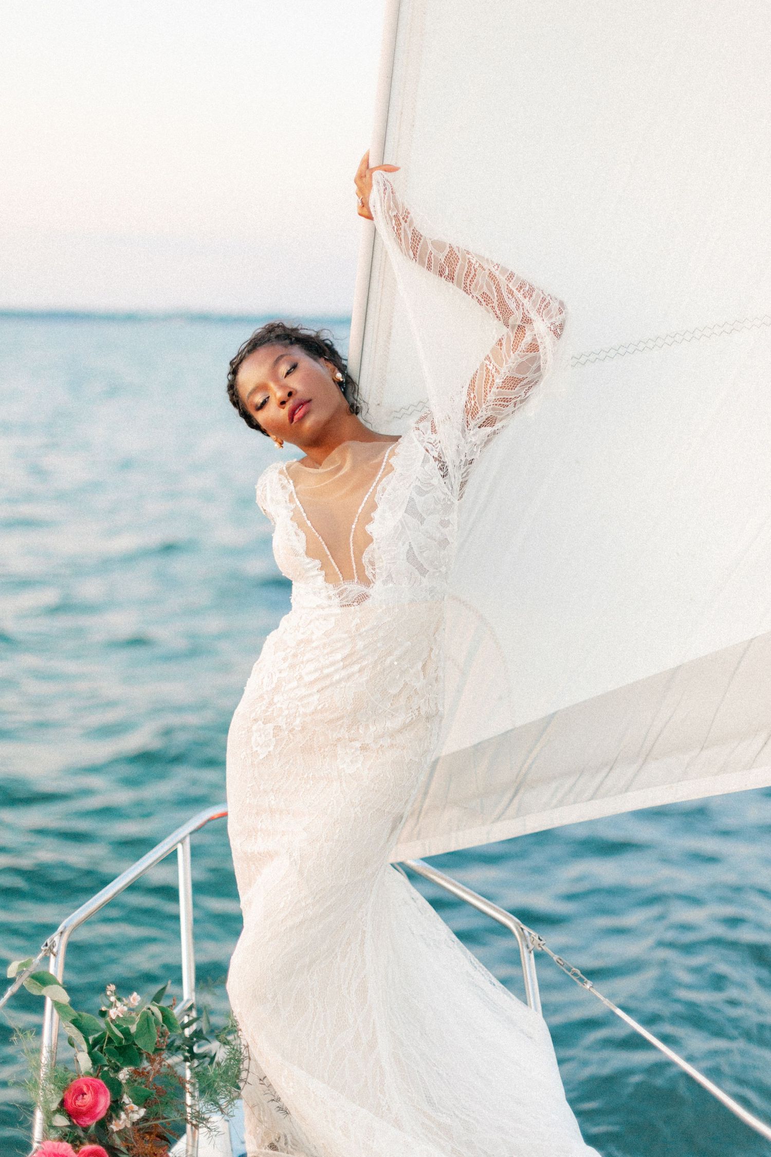 Bride in lace mermaid gown poses gracefully on sailboat deck with ocean backdrop.