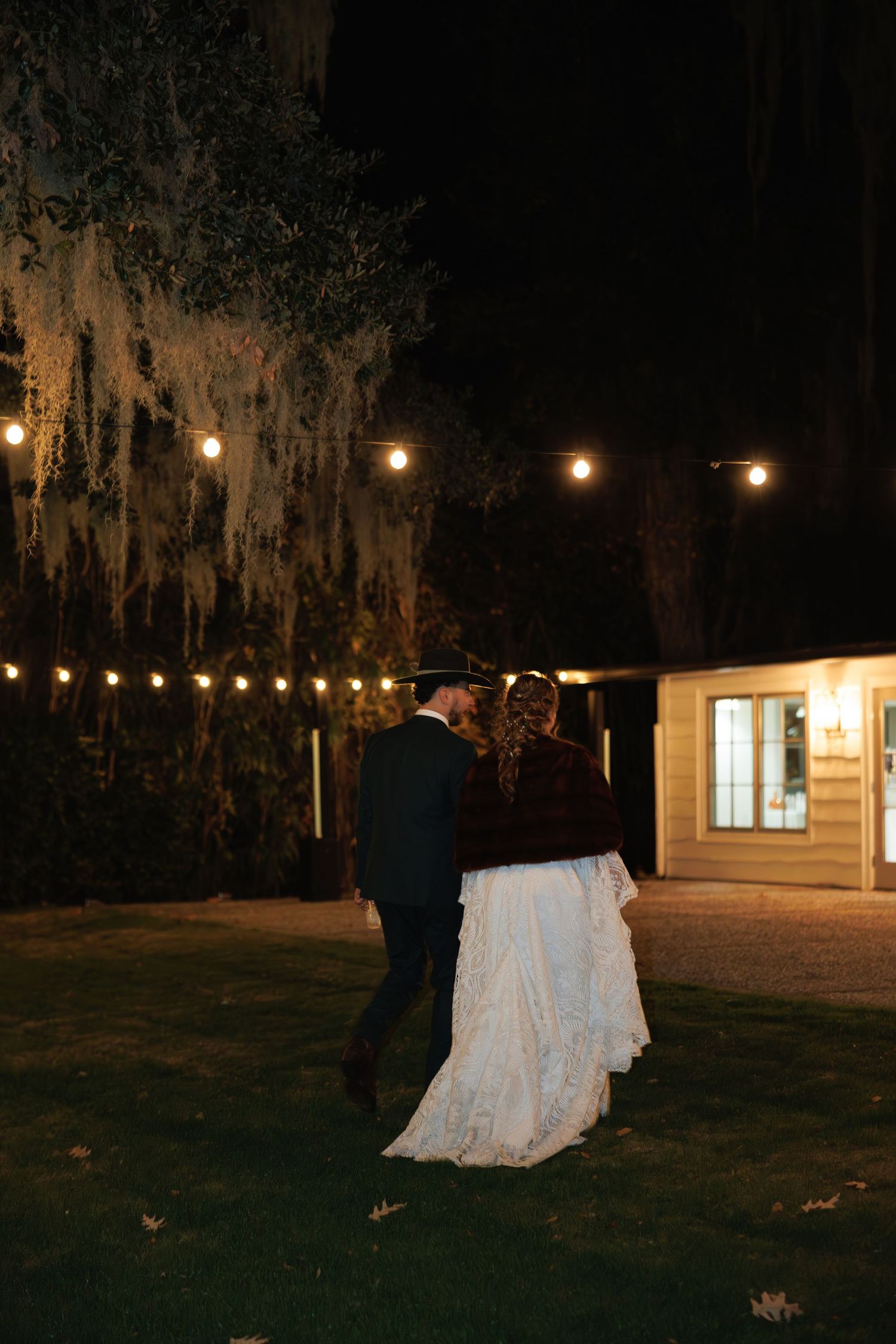Bride and groom walking together at night under Spanish moss trees with glowing string lights.