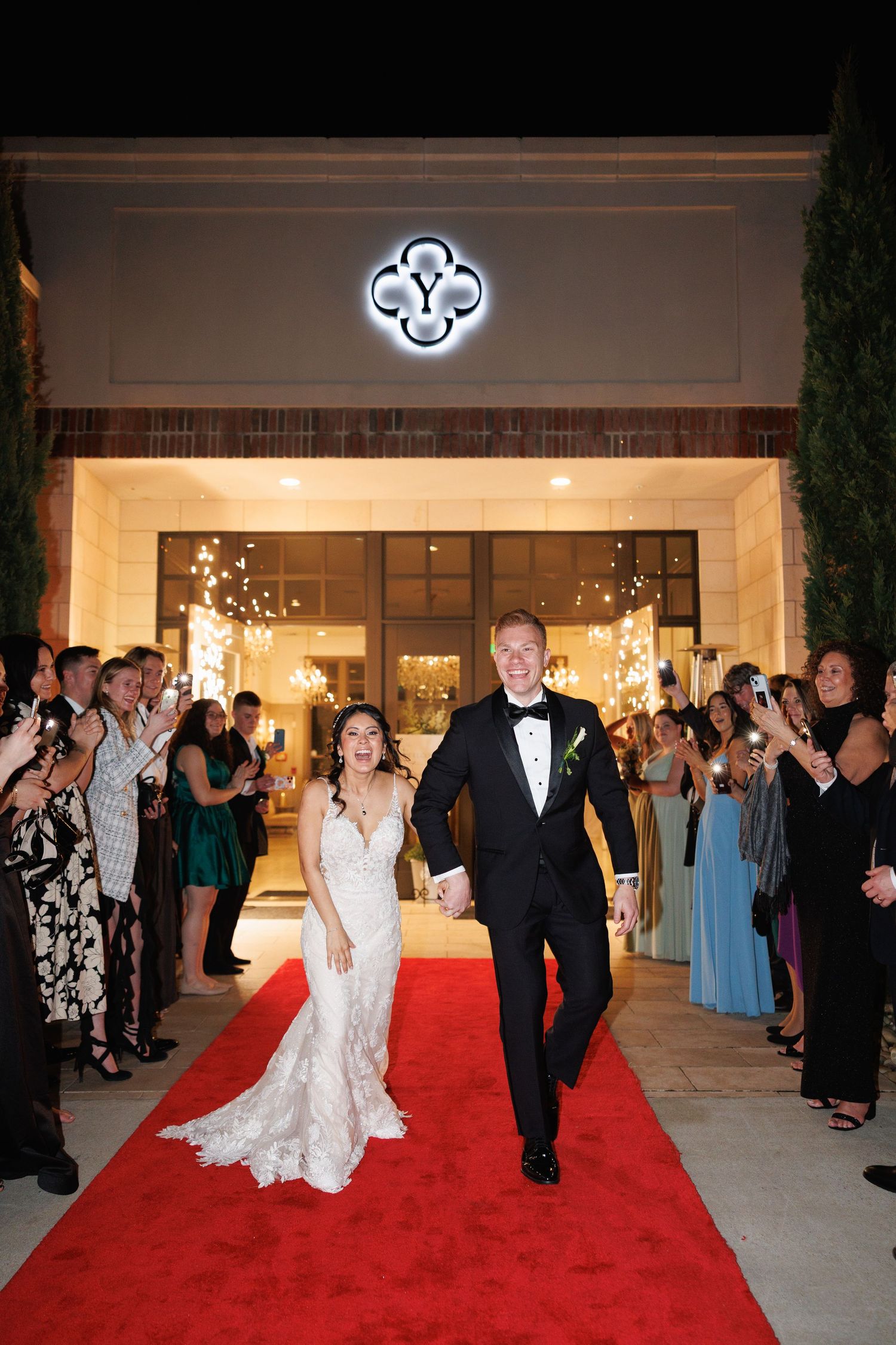 Newlyweds walk down a red carpet exit amid cheering guests outside an elegant wedding venue at night at the Yorkmont in Charlotte, NC.