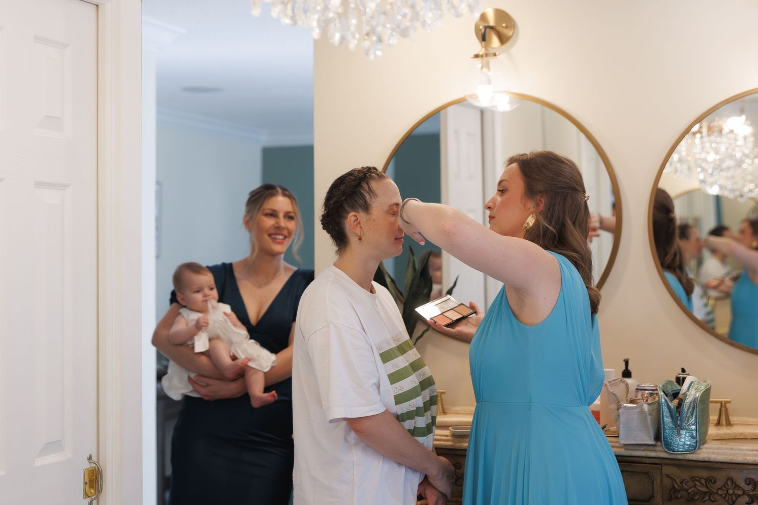 Woman in blue dress applies makeup to another person while a woman holding a baby watches in a bright bathroom.