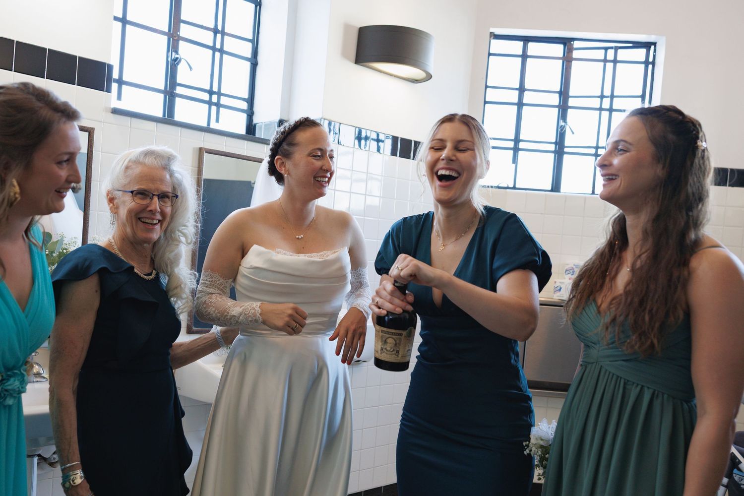 Bride and bridesmaids laughing while opening champagne bottle during wedding preparations.