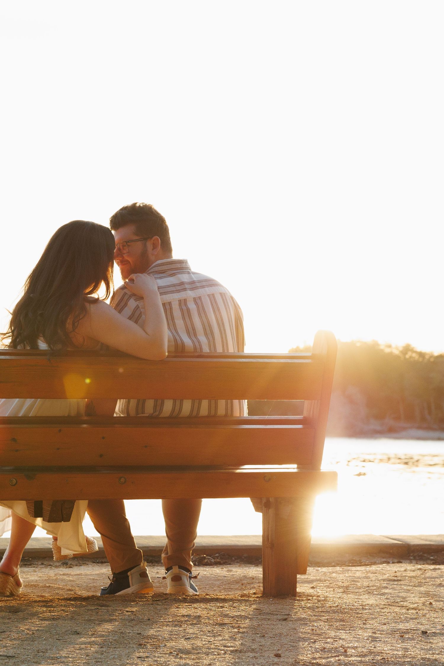 Couple sitting close together on a wooden bench by the water, bathed in warm golden sunset light.
