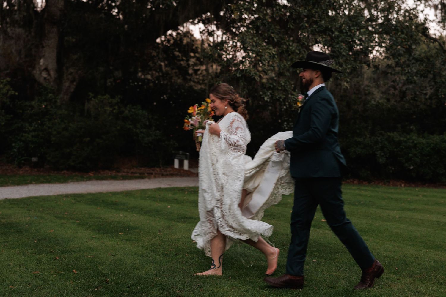 Bride in white lace gown walking barefoot with groom in green suit on garden lawn.