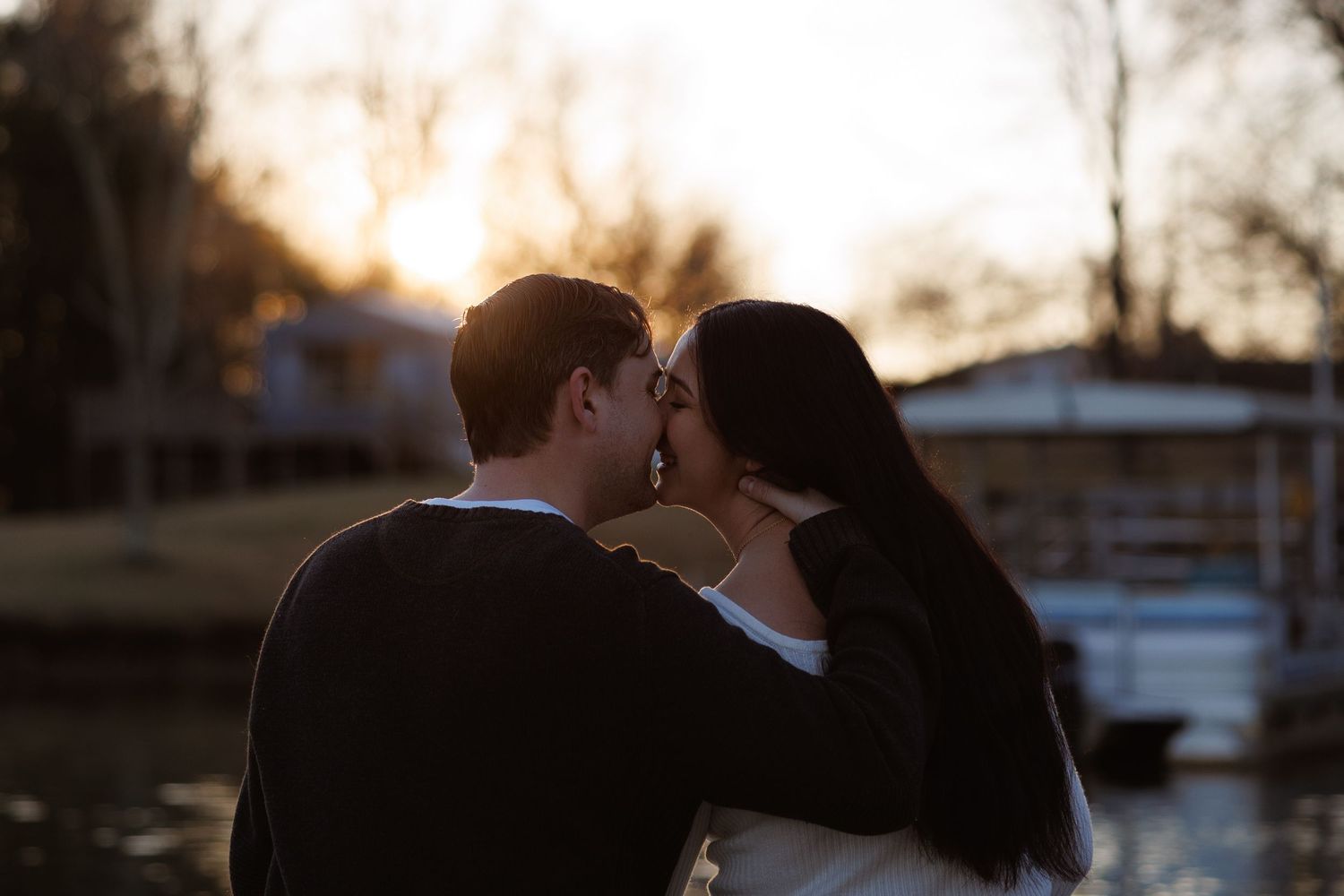 Couple sharing a romantic kiss at sunset by a lake, backlit by golden hour light.