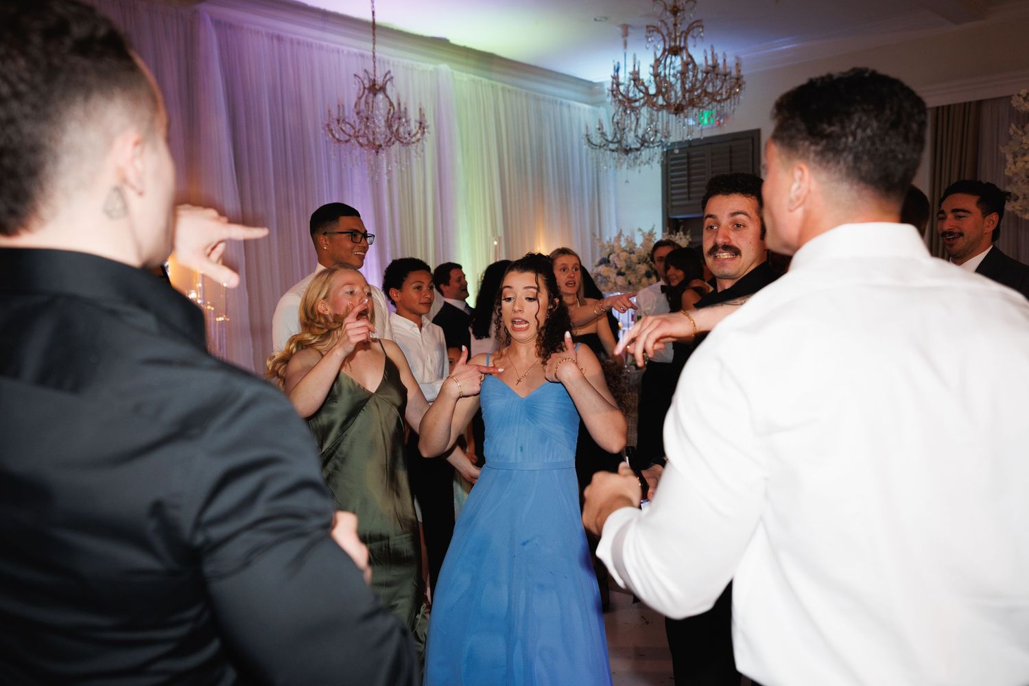 Guests in formal attire dance together on the floor at an elegant wedding reception with chandeliers.