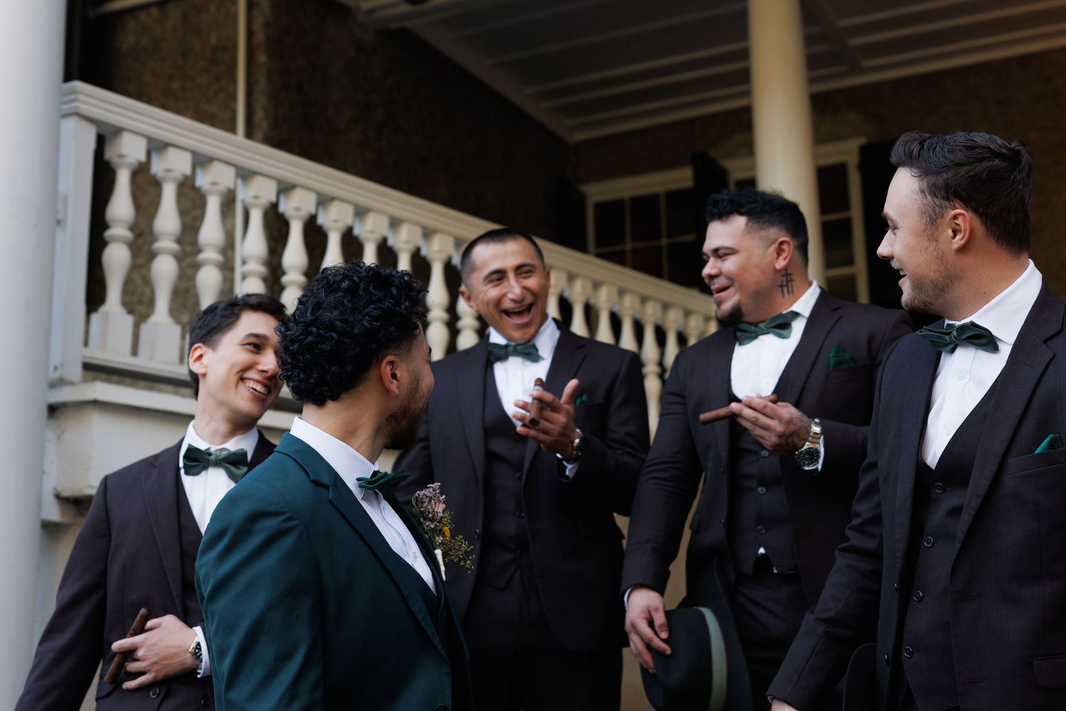 Groomsmen in dark suits and bow ties laughing together outside a venue with white balustrades.