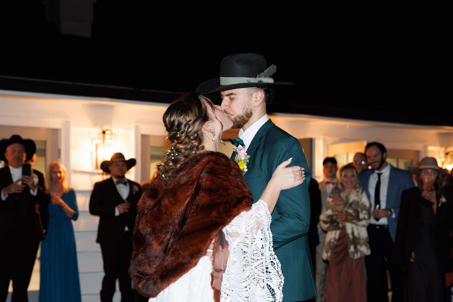 Newlyweds share first kiss at night wedding reception as guests celebrate in formal attire.