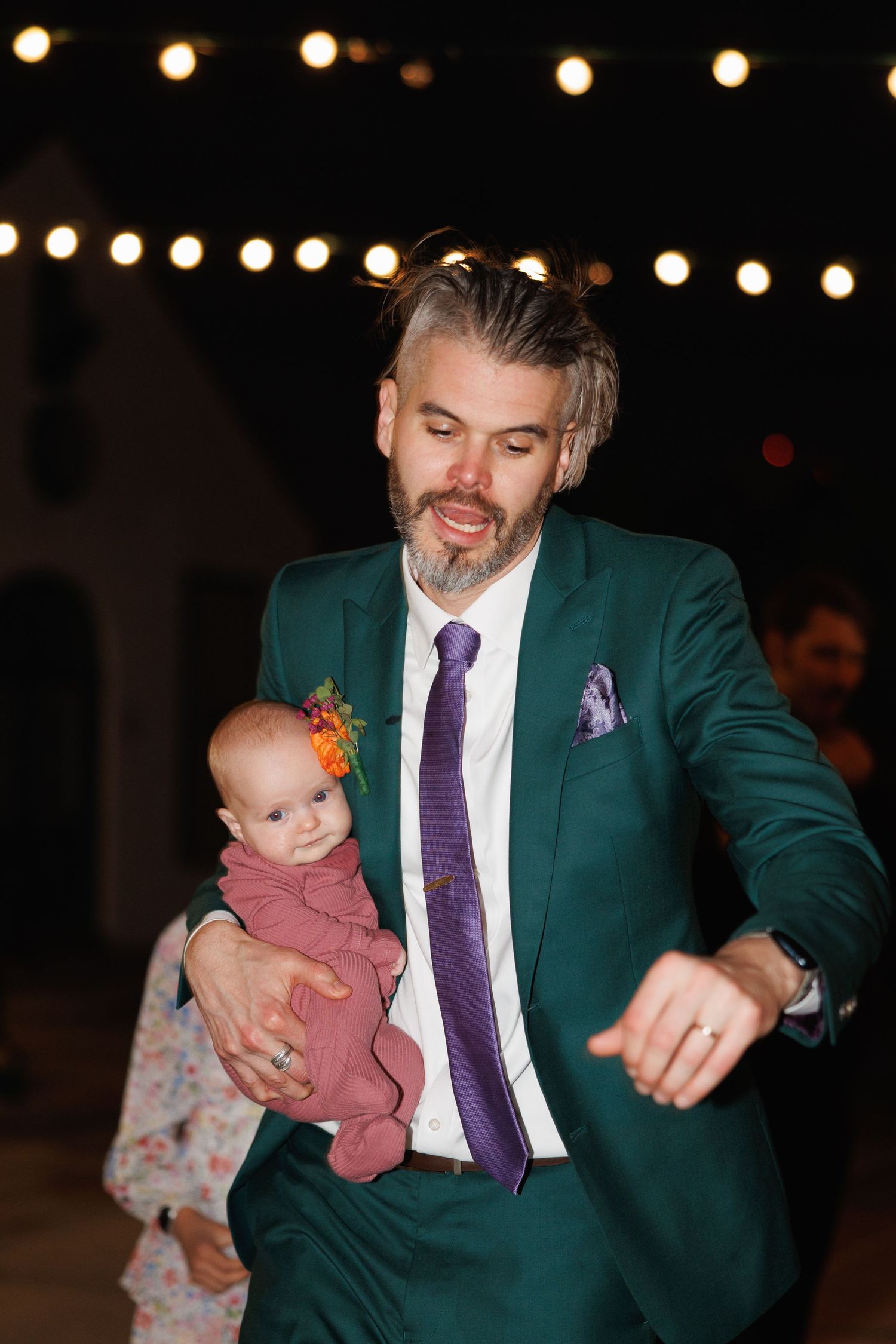 Man in green suit holding a baby at an outdoor evening wedding reception with string lights.
