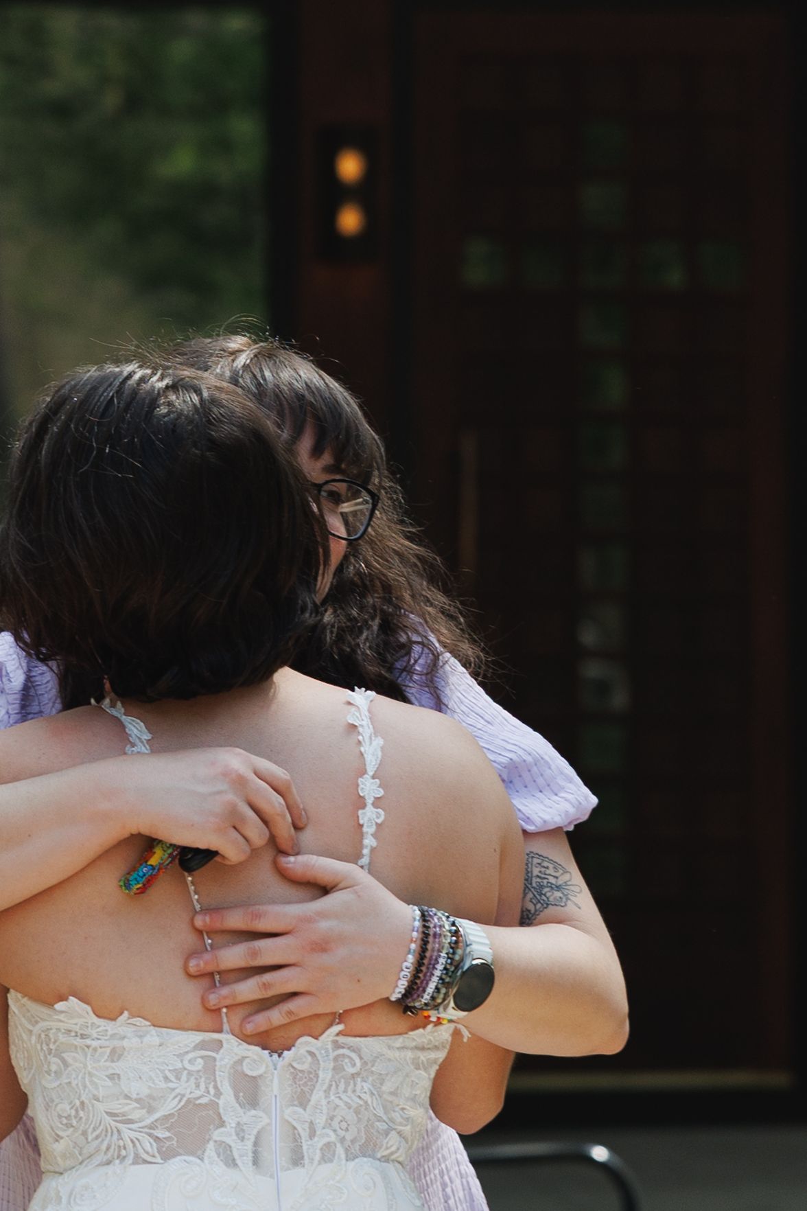 Two women share a warm embrace outdoors, one wearing a white lace dress with a tattoo visible on her arm.